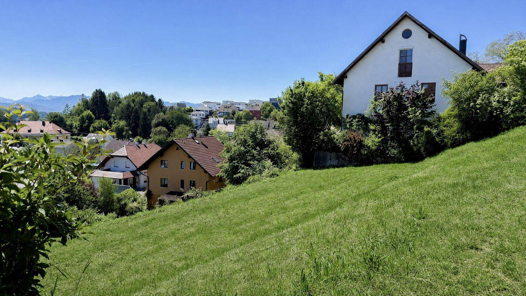 Blick auf Wohnhäuser an einem grünen Hang mit weitem Blick auf die umliegende Landschaft und Berge.
