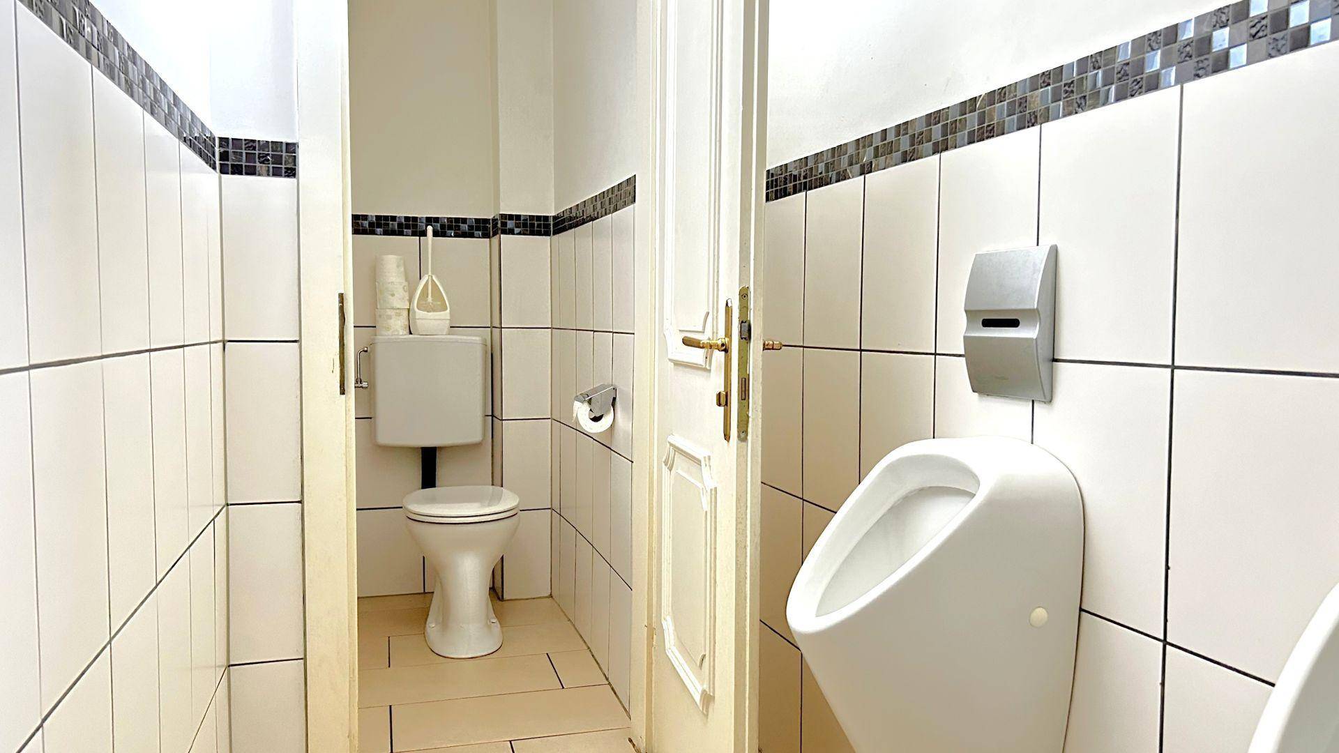 Toilettenraum mit weißen Fliesen, Urinal und WC, getrennt durch eine Tür.