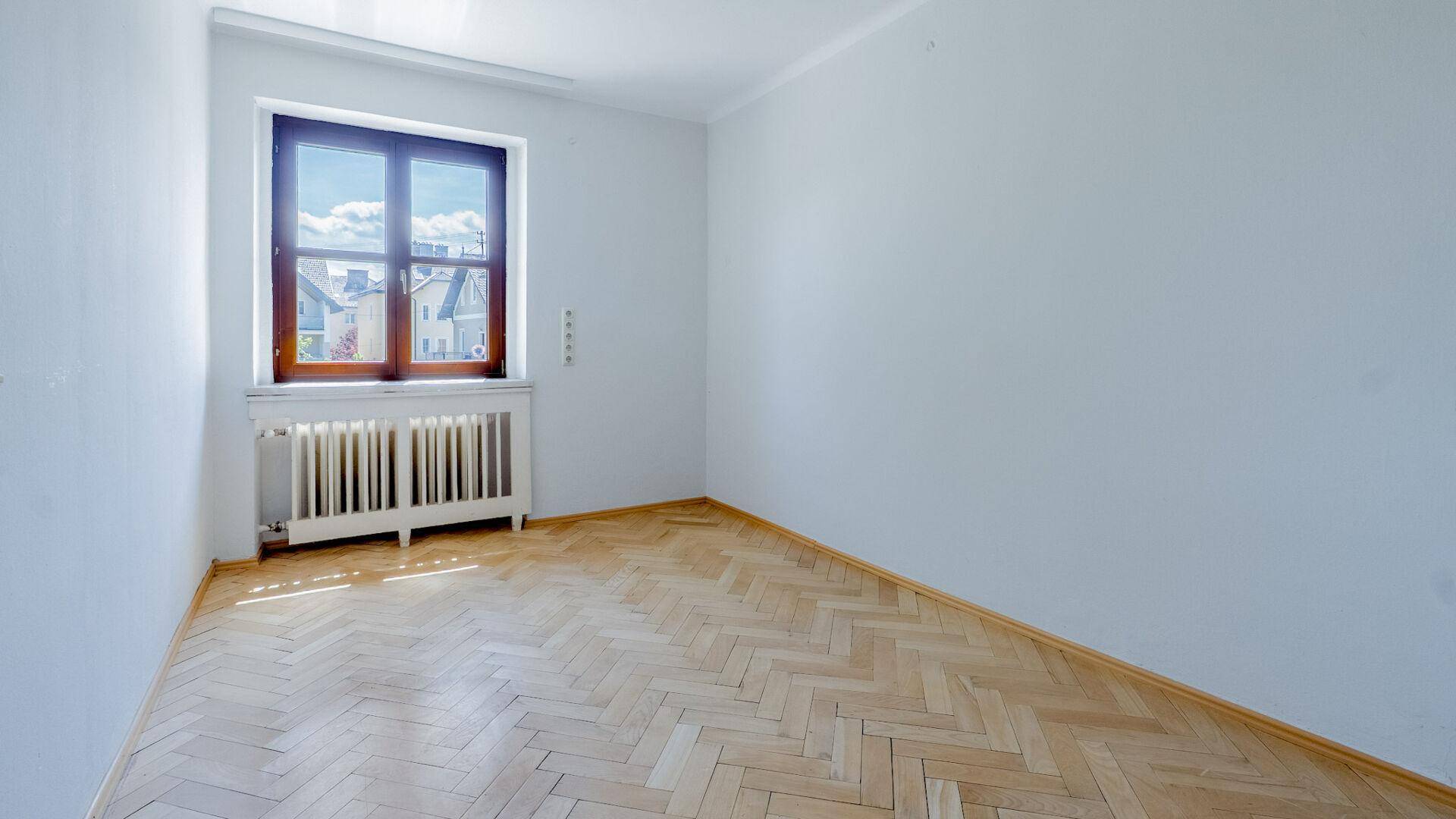 Helles leeres Zimmer mit Fischgrätparkett und Fenster, das viel Tageslicht bietet.