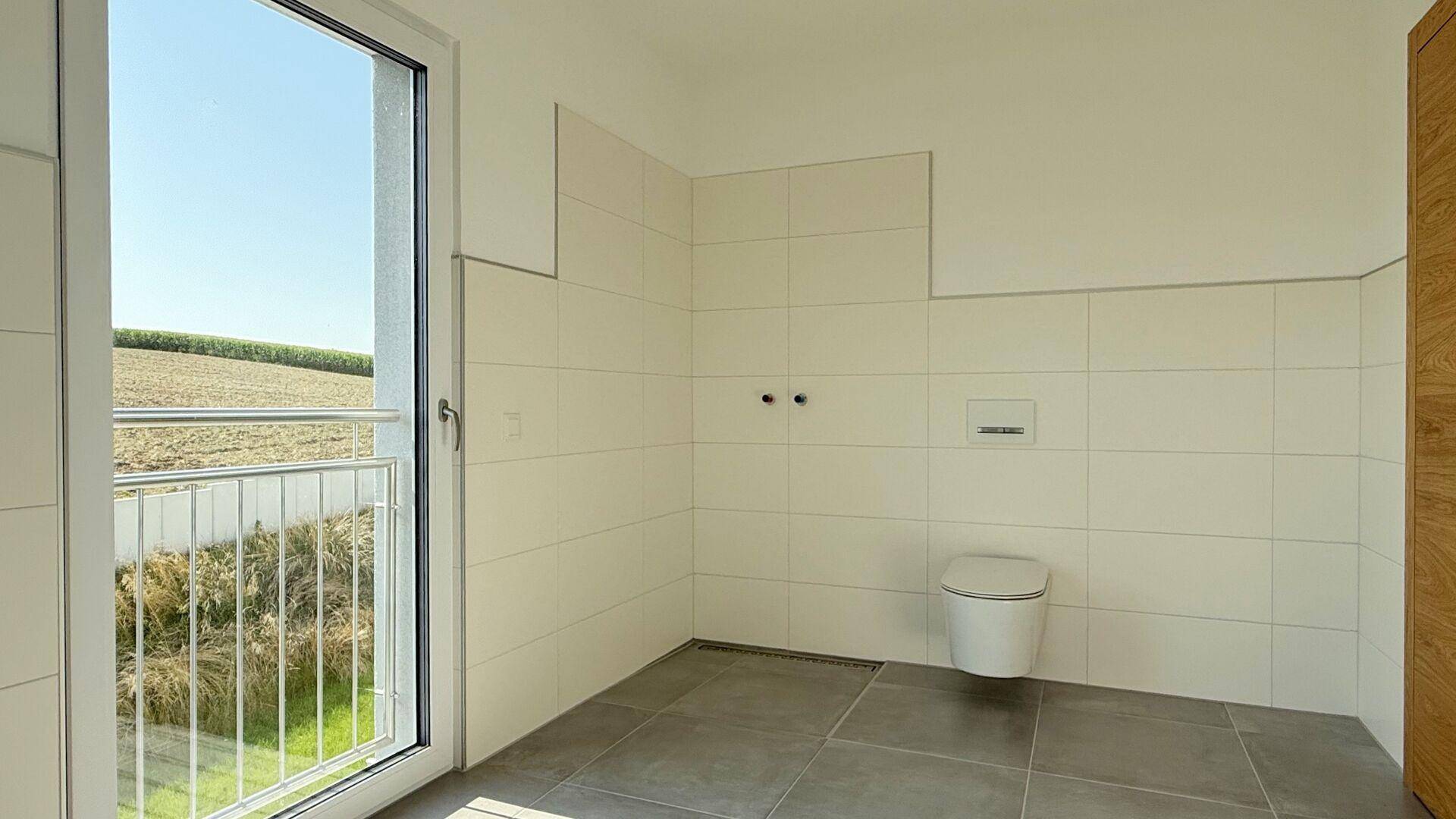 Unfertiges Badezimmer mit Wand-WC, Fliesenboden und französischem Balkon mit Blick ins Grüne.