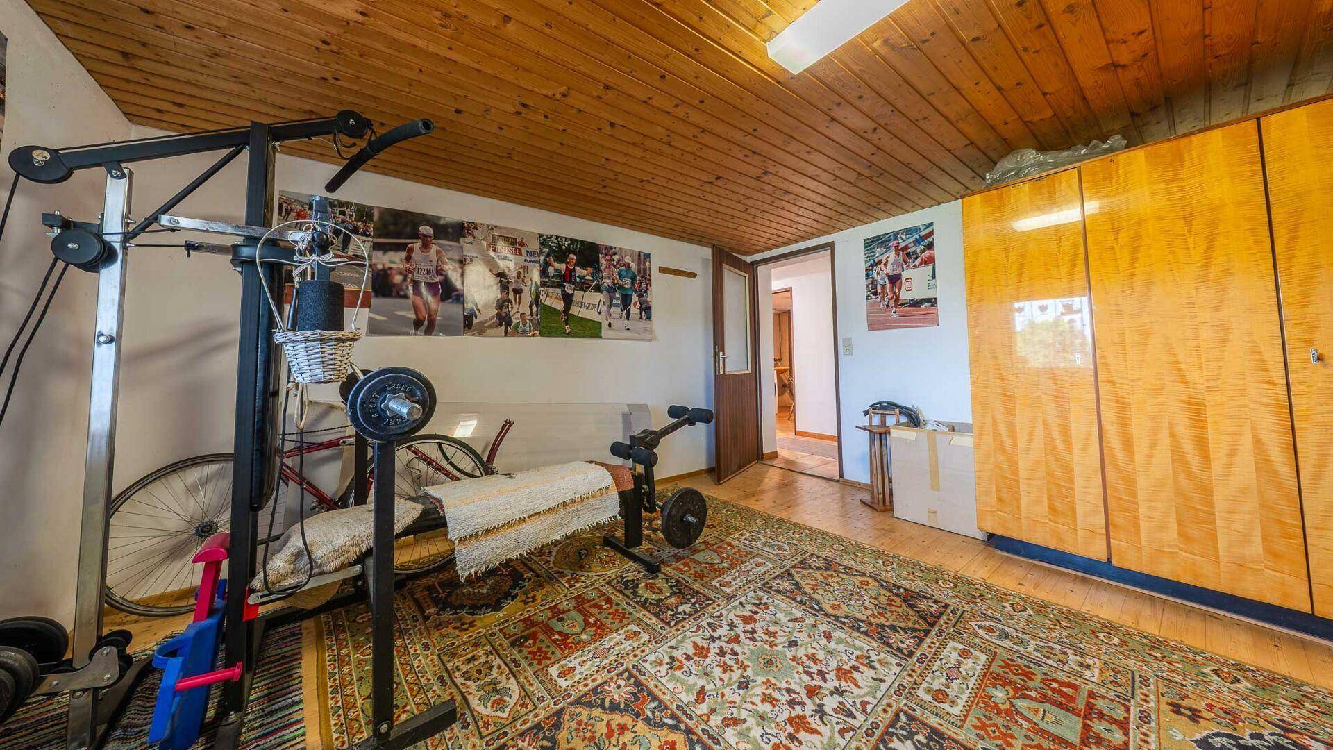 Hobbyraum mit Fitnessgeräten, großem Kleiderschrank und gemusterter Teppichboden, unter einer Holzdecke.