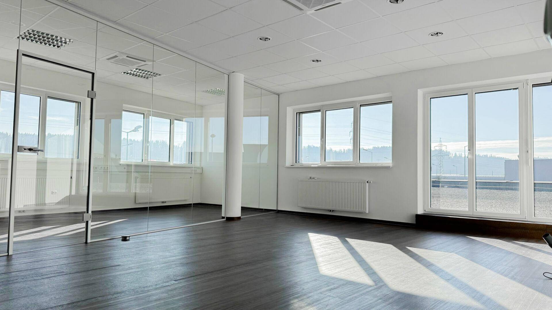 Moderner Büroraum mit Glaswänden, großen Fenstern und dunklem Boden, bietet eine helle Arbeitsatmosphäre.