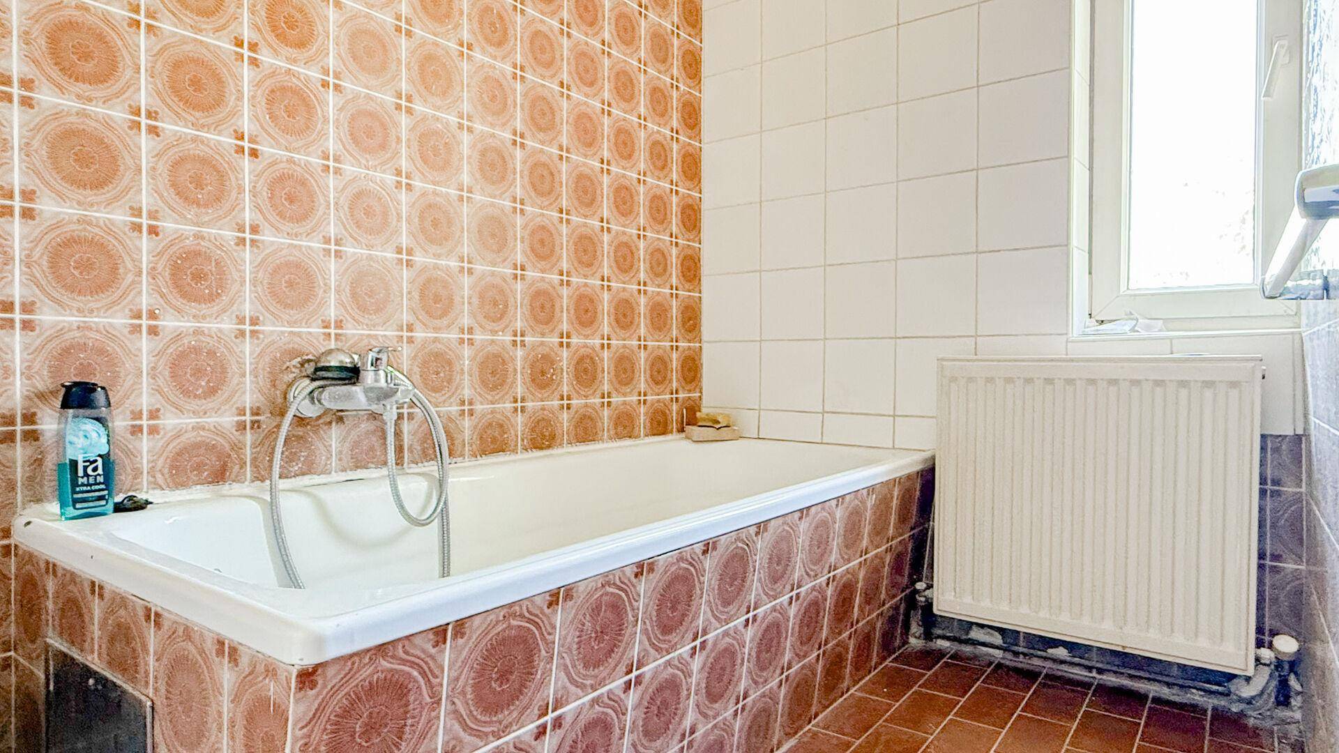 Badezimmer mit Badewanne, auffälligen orange-braunen Fliesen und einem Heizkörper unter dem Fenster.