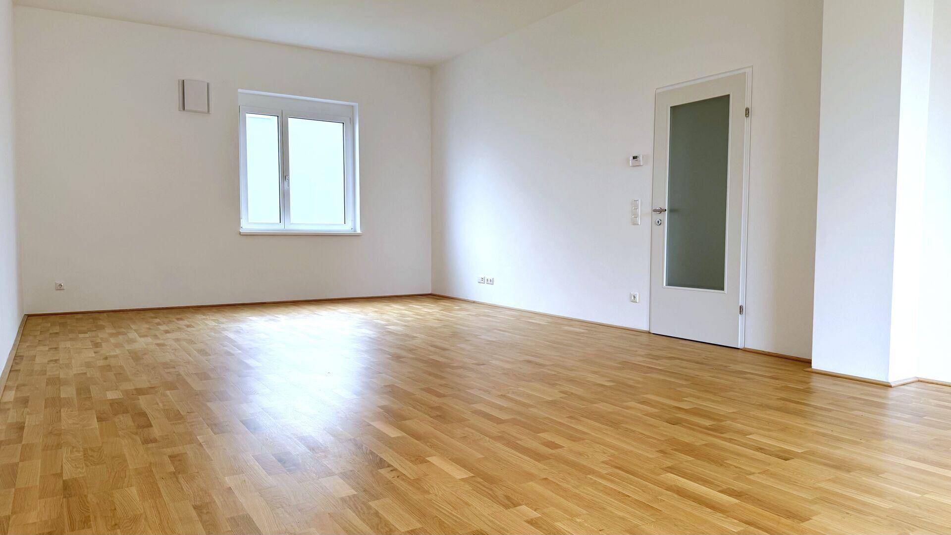 Geräumiges, helles Zimmer mit Parkettboden und einem Fenster, das viel Tageslicht hereinlässt.