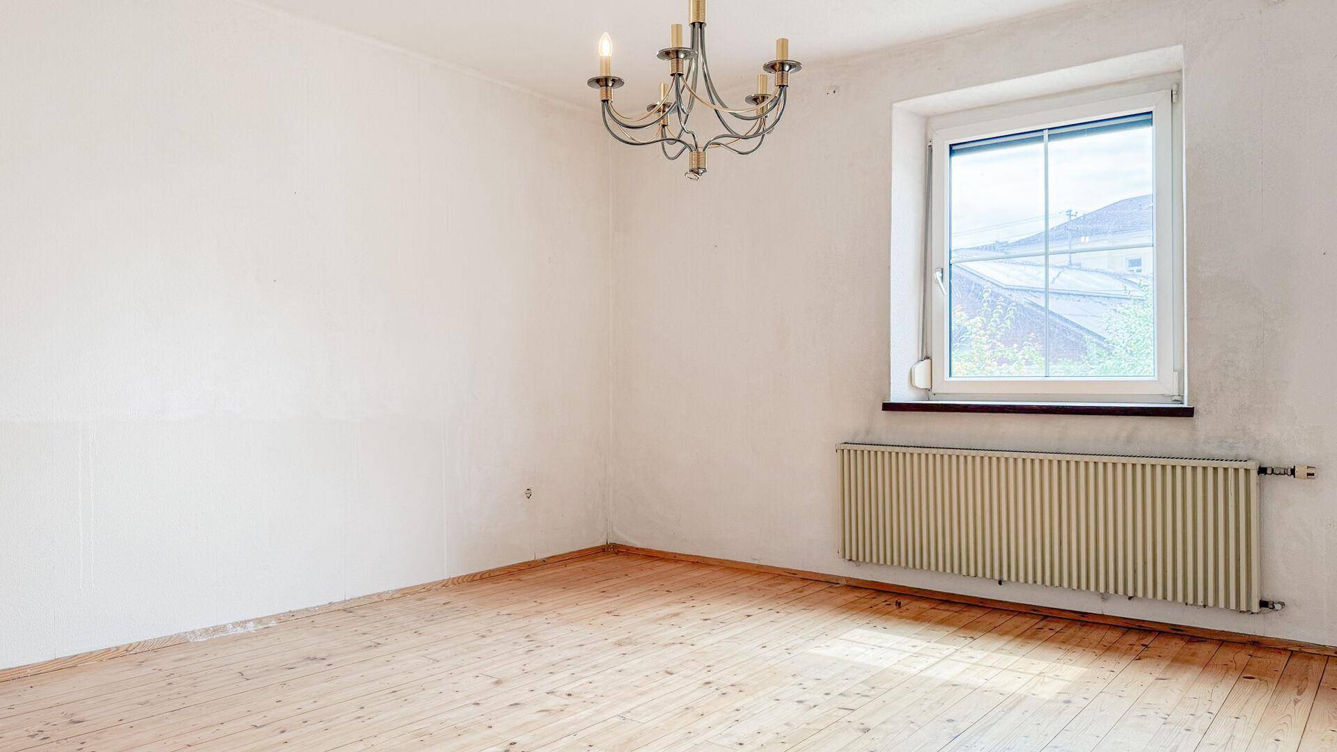Leeres Zimmer mit Holzdielenboden, Fenster und einem klassischen Kronleuchter an der Decke.