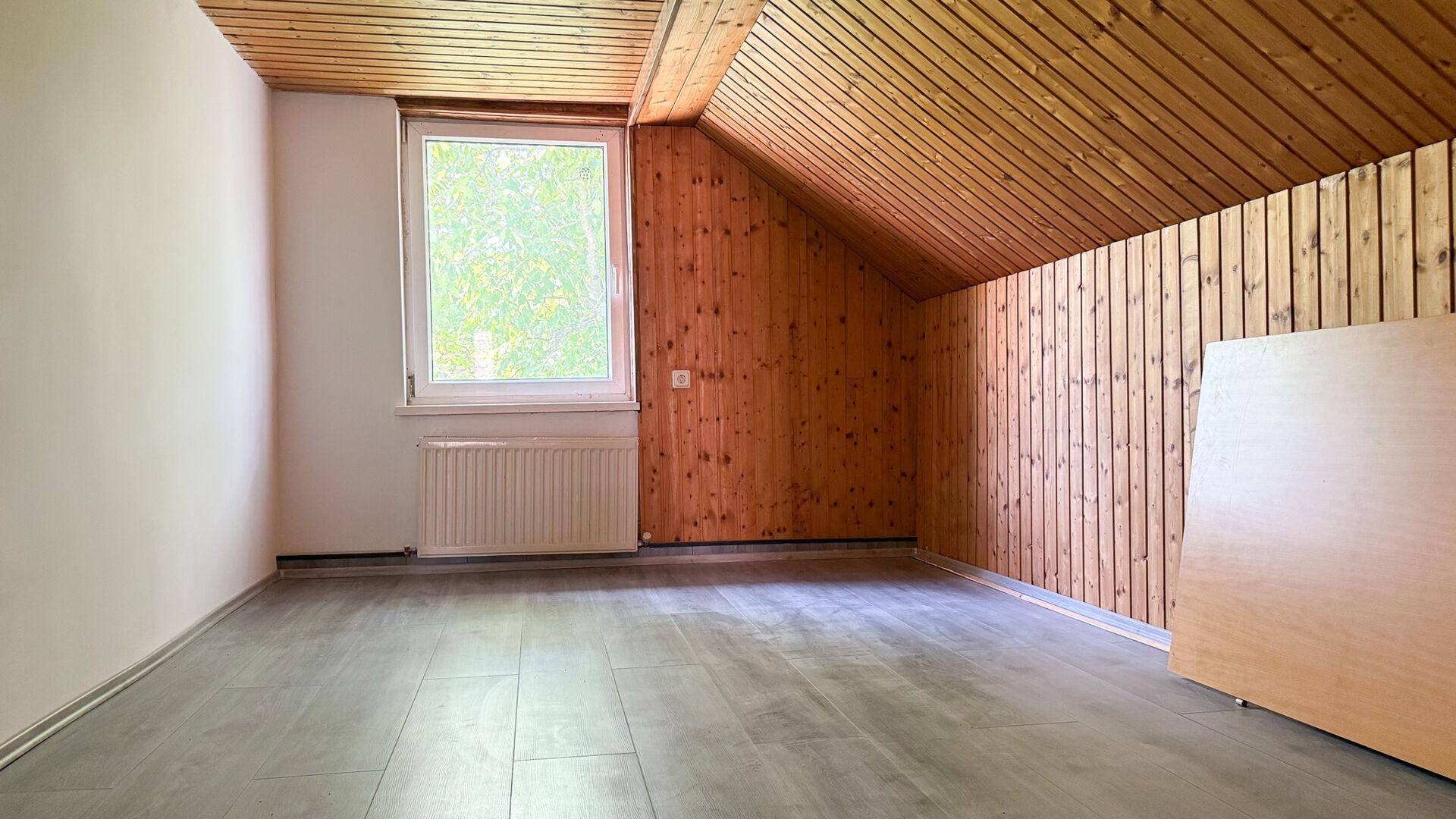 Leeres Dachzimmer mit Holzverkleidung, schräger Decke, Fenster und Heizkörper.