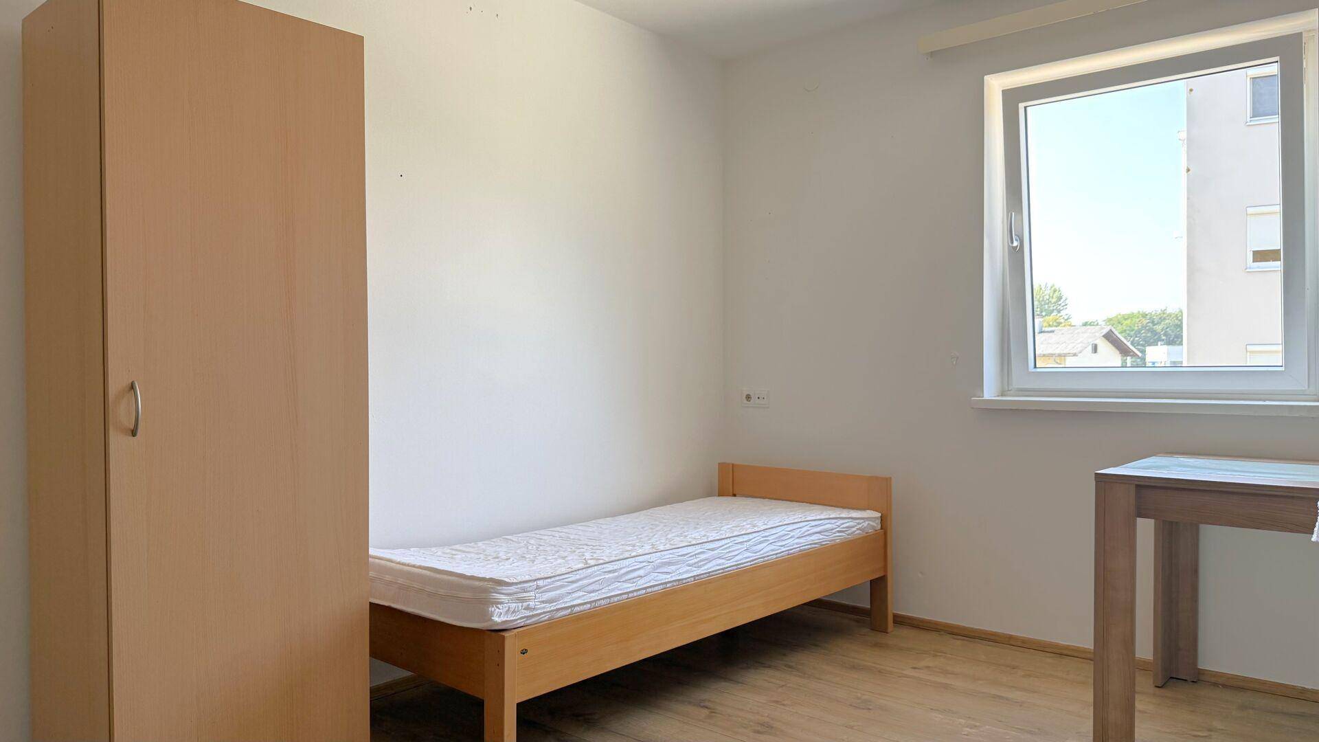 Einfaches Schlafzimmer mit Einzelbett, Kleiderschrank und Fenster.