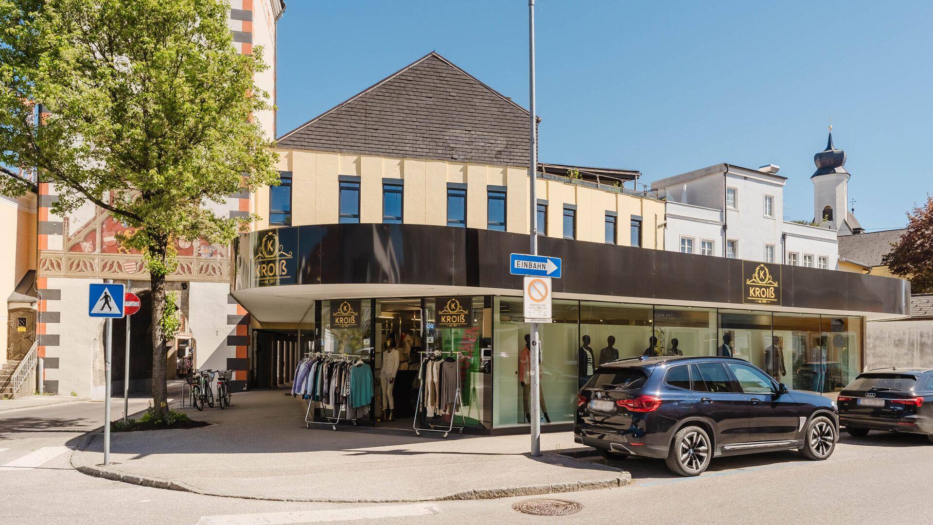 Geschäftshaus mit dunkler Fassade und großen Schaufenstern an einer belebten Straße mit parkenden Autos.