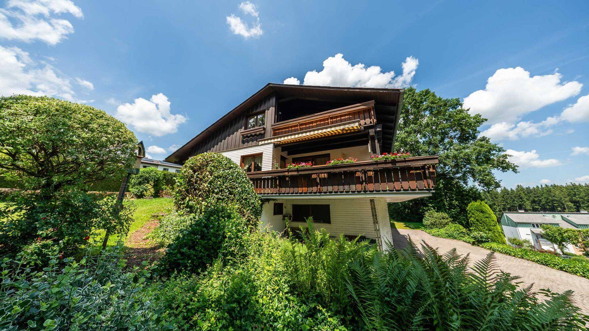 Traditionelles Haus mit Holzbalkon und Blumenkästen, umgeben von üppigem Grün unter blauem Himmel.