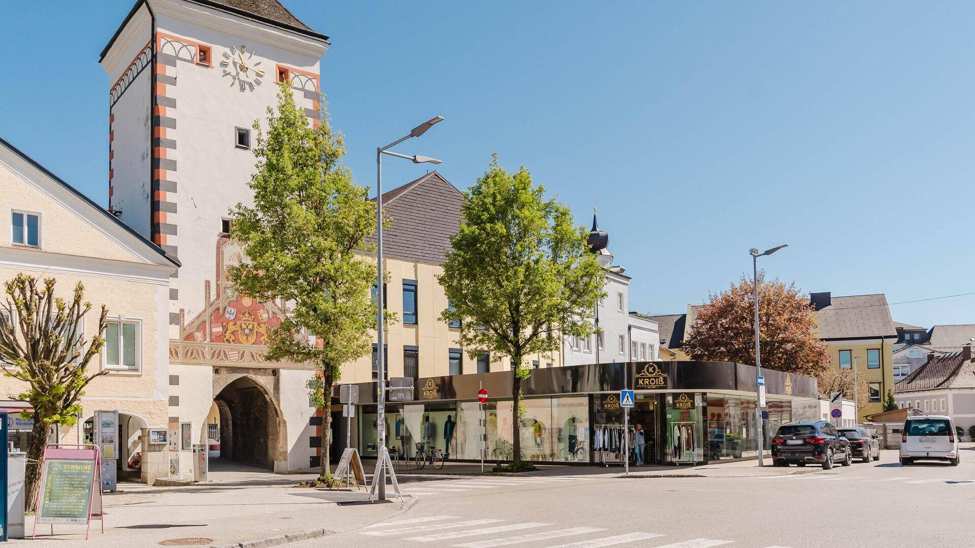 Belebtes Stadtbild mit einem Uhrturm, einem modernen Geschäft und parkenden Autos.