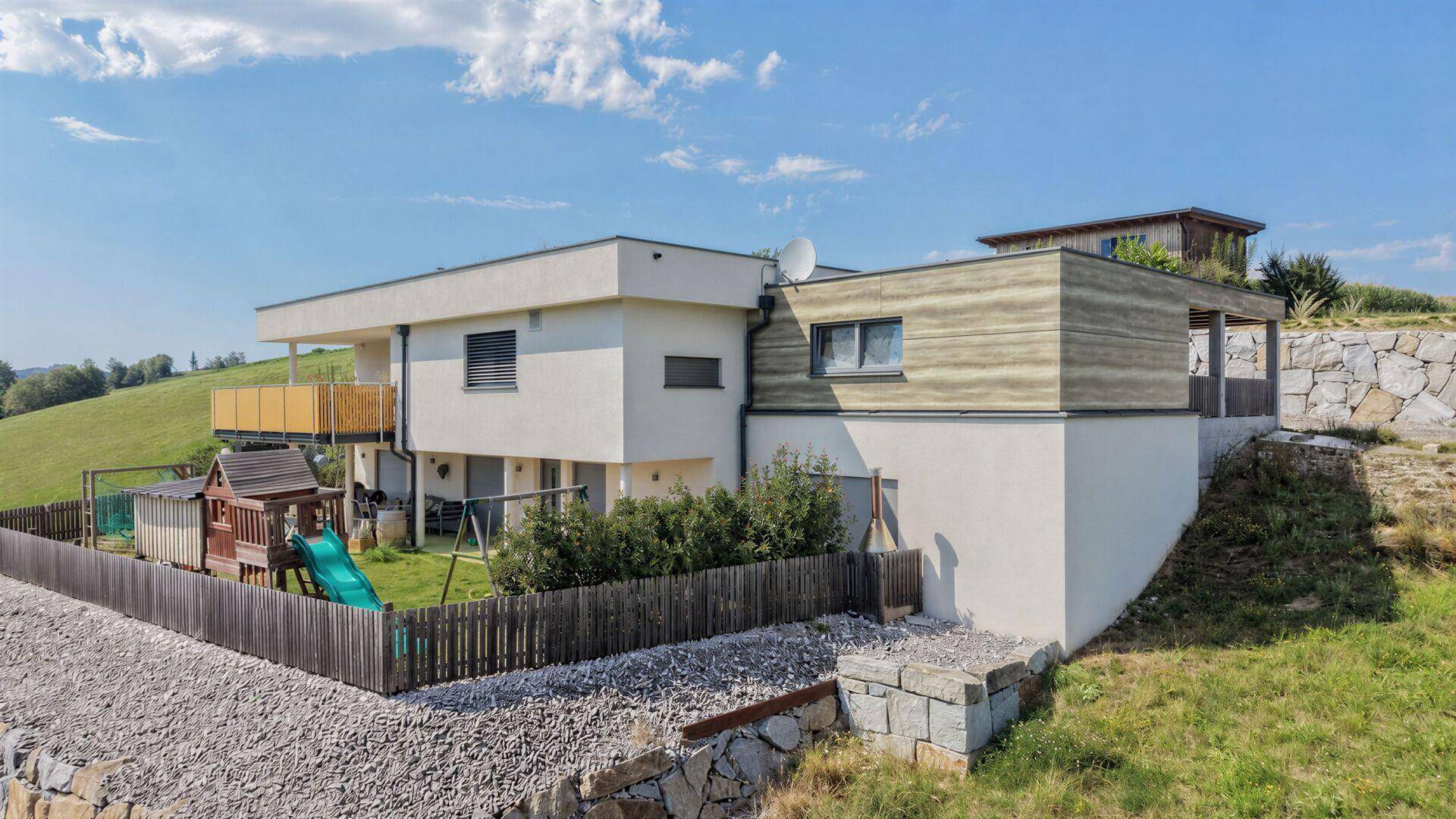 Seitenansicht des Hauses mit Spielplatz im Garten und moderner Architektur in ländlicher Umgebung.