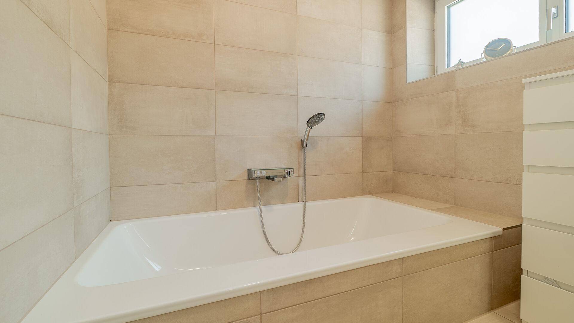 Modernes Badezimmer mit beigefarbenen Fliesen, integrierter Badewanne und Fenster für Tageslicht.