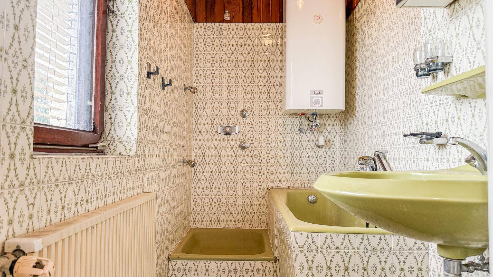 Badezimmer mit gemusterten Fliesen, kleiner Badewanne, grünem Waschbecken und Warmwasserbereiter.
