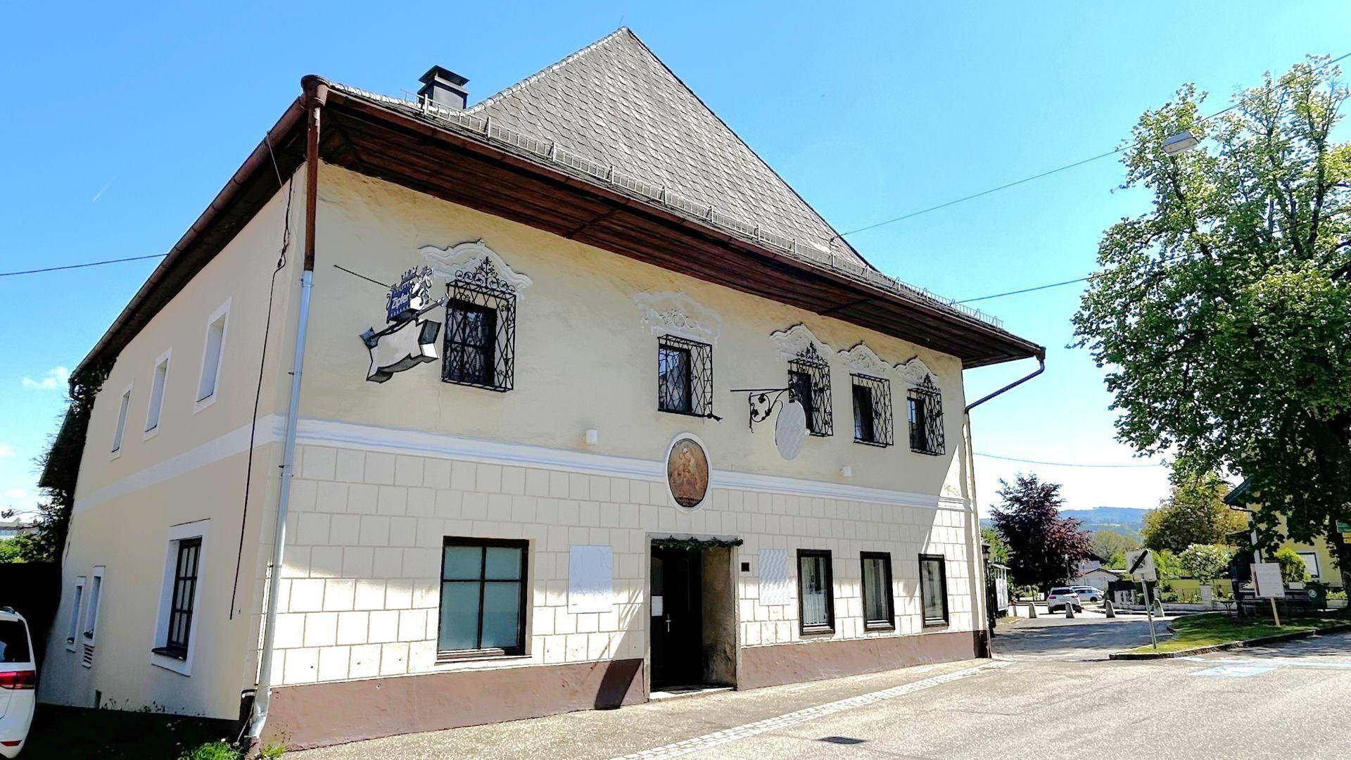 Traditionelles Gebäude mit bemalter Fassade, Satteldach und mehreren Fenstern in ländlicher Umgebung.