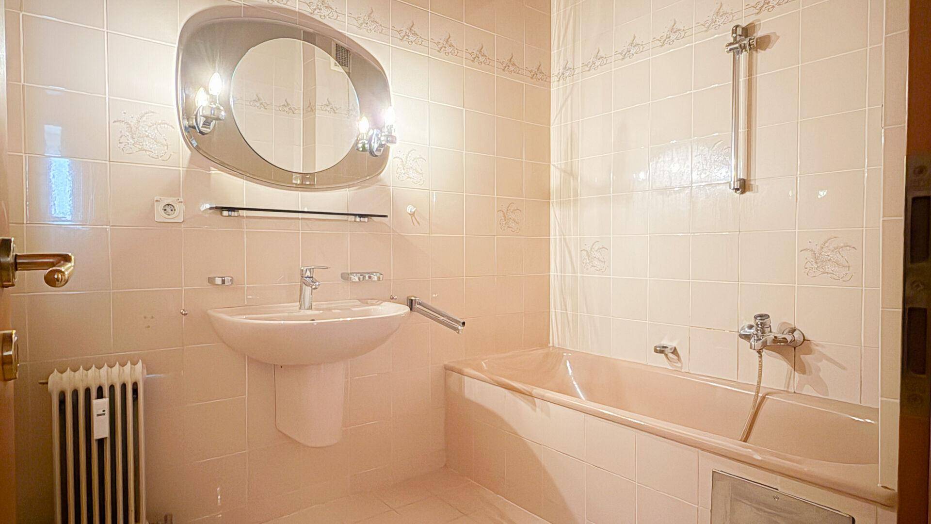 Badezimmer mit beigefarbenen Fliesen, Badewanne, Waschbecken und einem Spiegel mit Beleuchtung.
