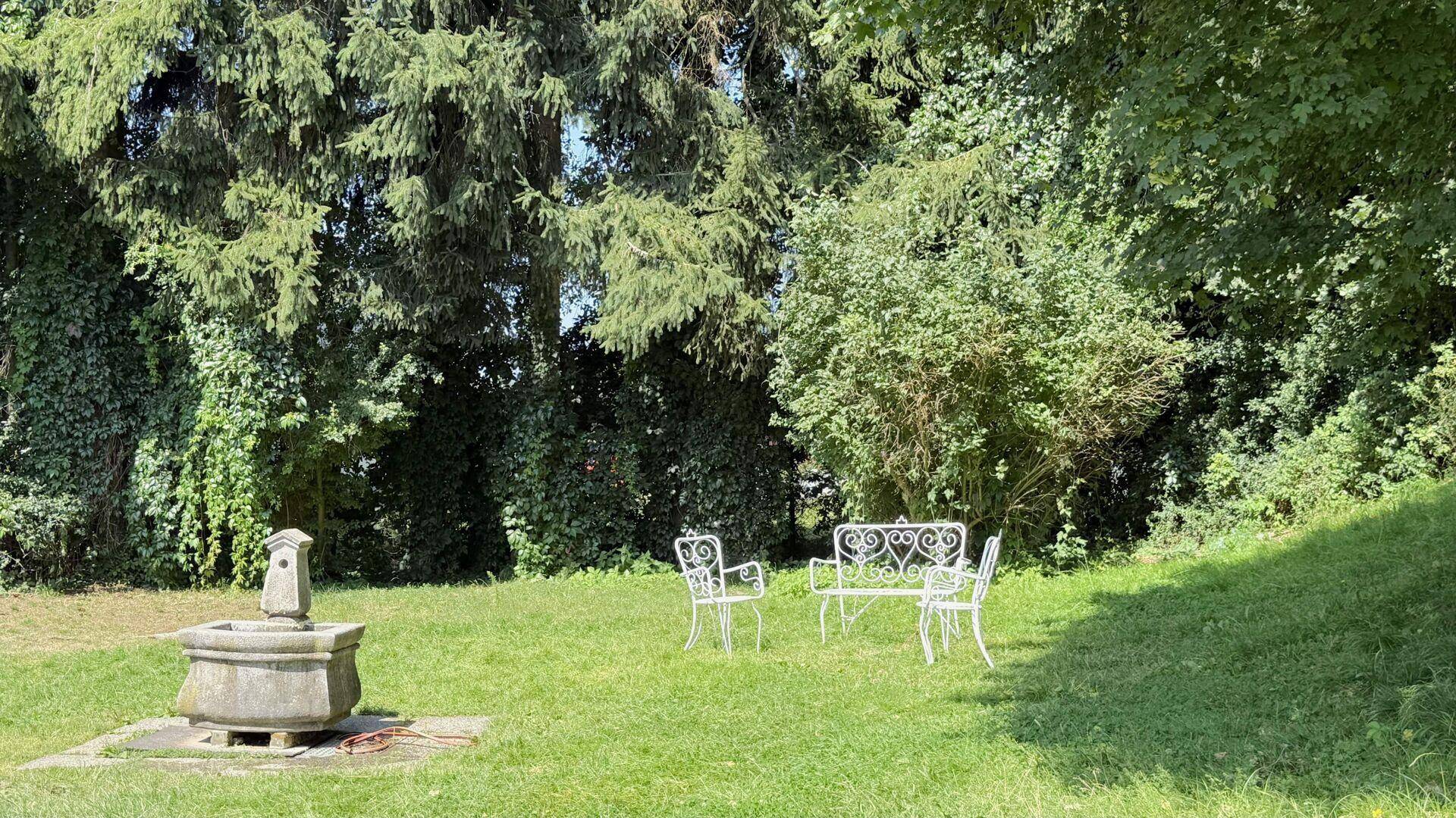 Gepflegter Gartenbereich mit einem Steinbrunnen und weißen Gartenmöbeln unter hohen Bäumen.