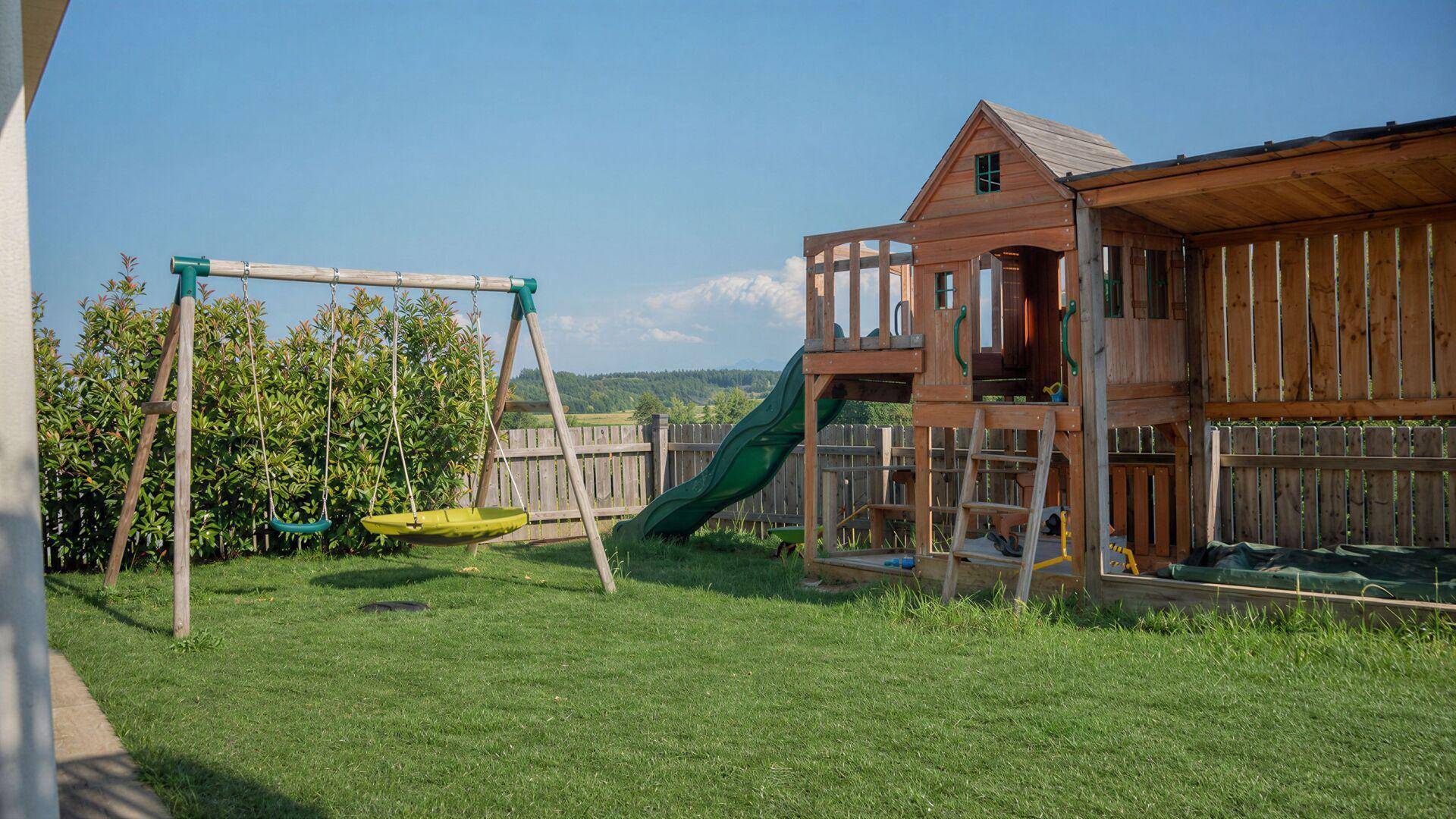 Großer Garten mit Spielplatz, Schaukel und Rutsche, umgeben von einem Holzzaun.