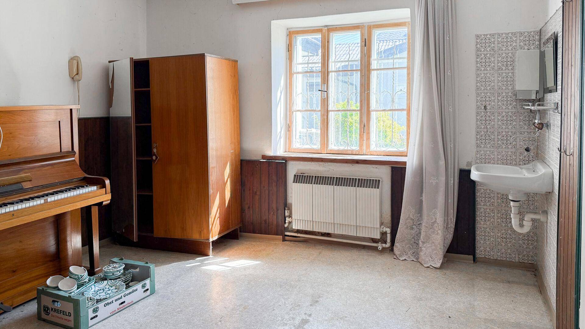 Raum mit Klavier, Schrank und kleinem Waschbecken, ausgestattet mit einem Fenster und Heizkörper.