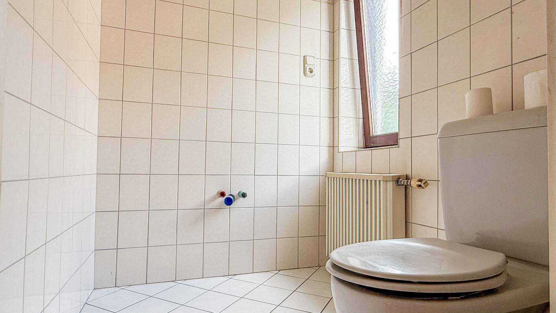 Separates WC mit weißen Fliesen, Toilette und einem Fenster für natürliche Belüftung.