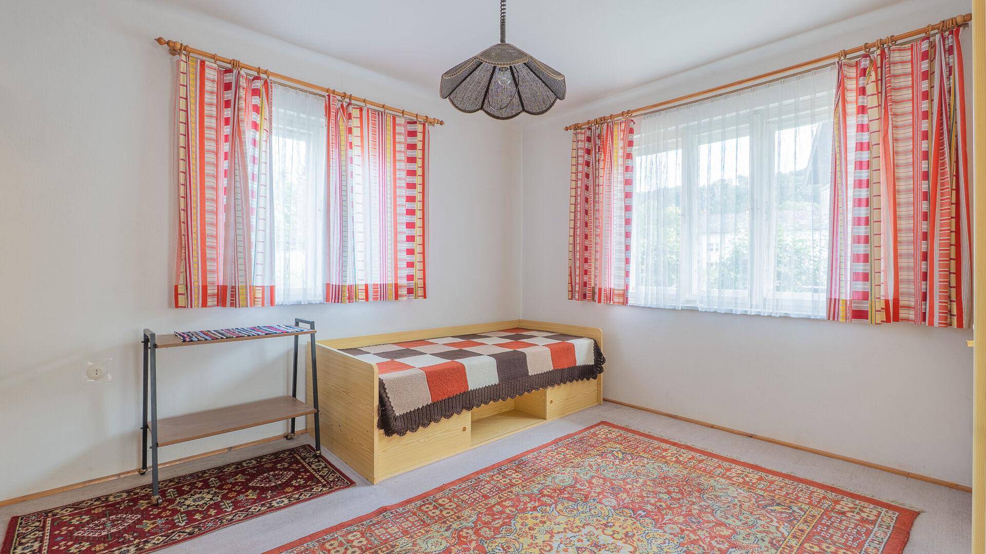 Schlafzimmer mit Einzelbett, Teppich und bunten Vorhängen, hell und freundlich.