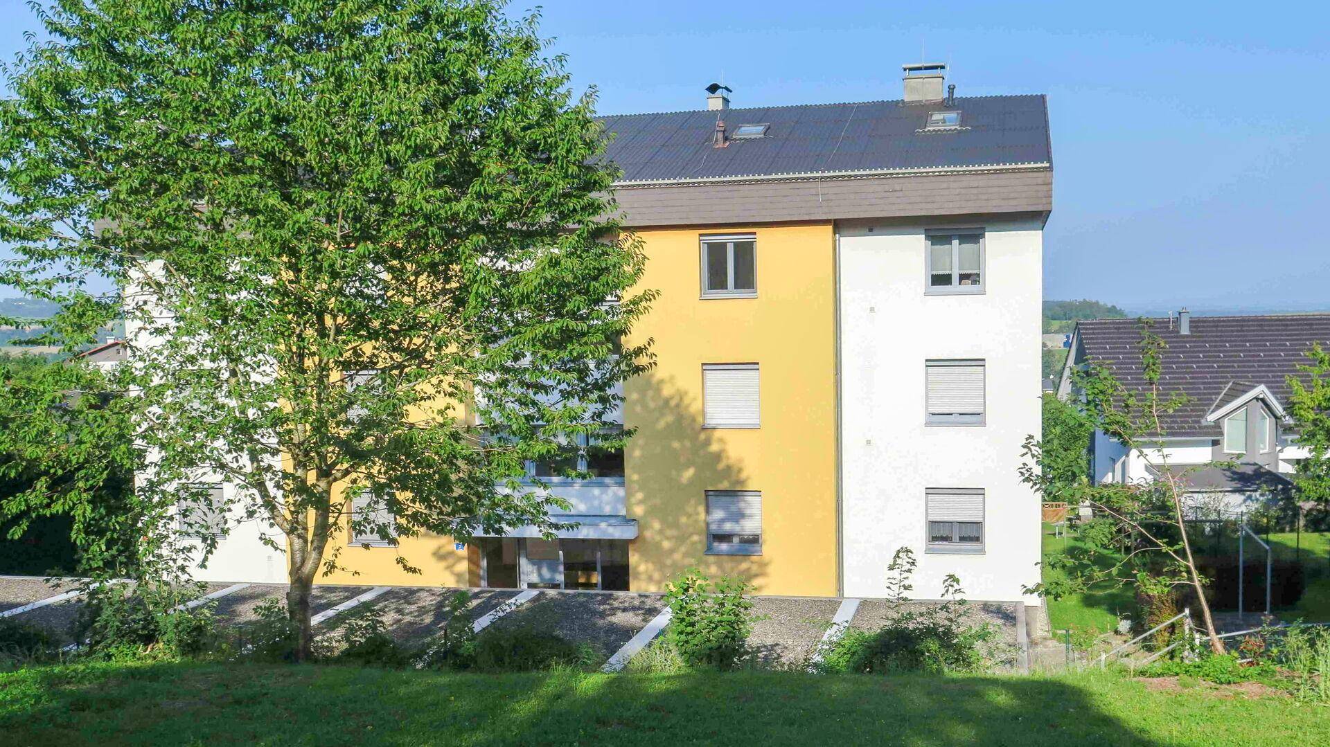 Rückseite eines Mehrfamilienhauses mit Bäumen und grüner Umgebung.