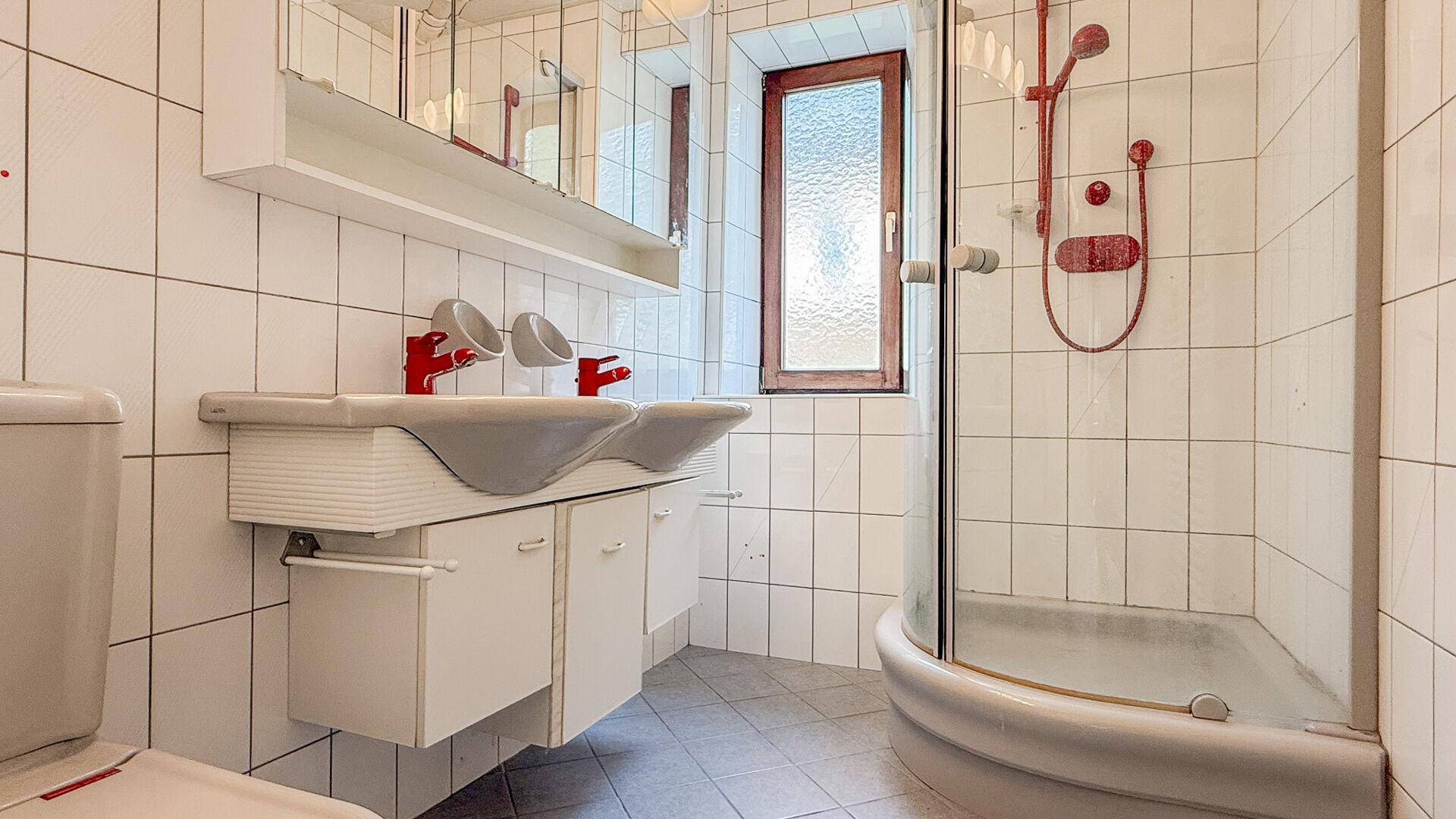 Badezimmer mit weißen Fliesen, Duschkabine, Toilette und Doppelwaschbecken mit roten Akzenten.