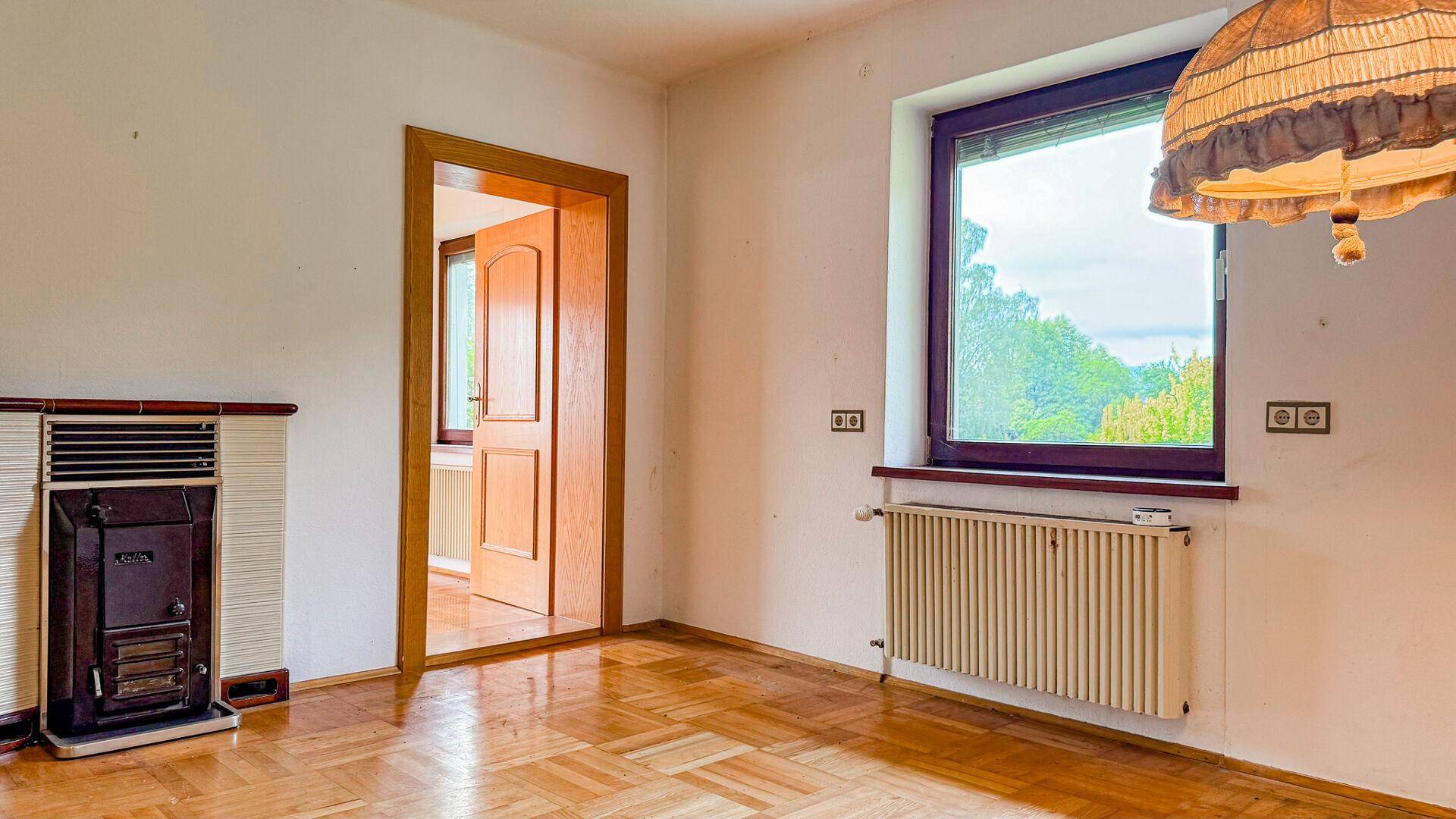 Helle Ecke eines Zimmers mit Parkettboden, Fenster, Heizkörper und einem traditionellen Ofen.