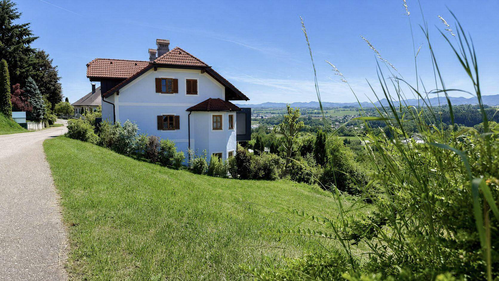 Ein weißes Haus mit rotem Ziegeldach an einem Hang, umgeben von grünen Wiesen und mit Fernblick.
