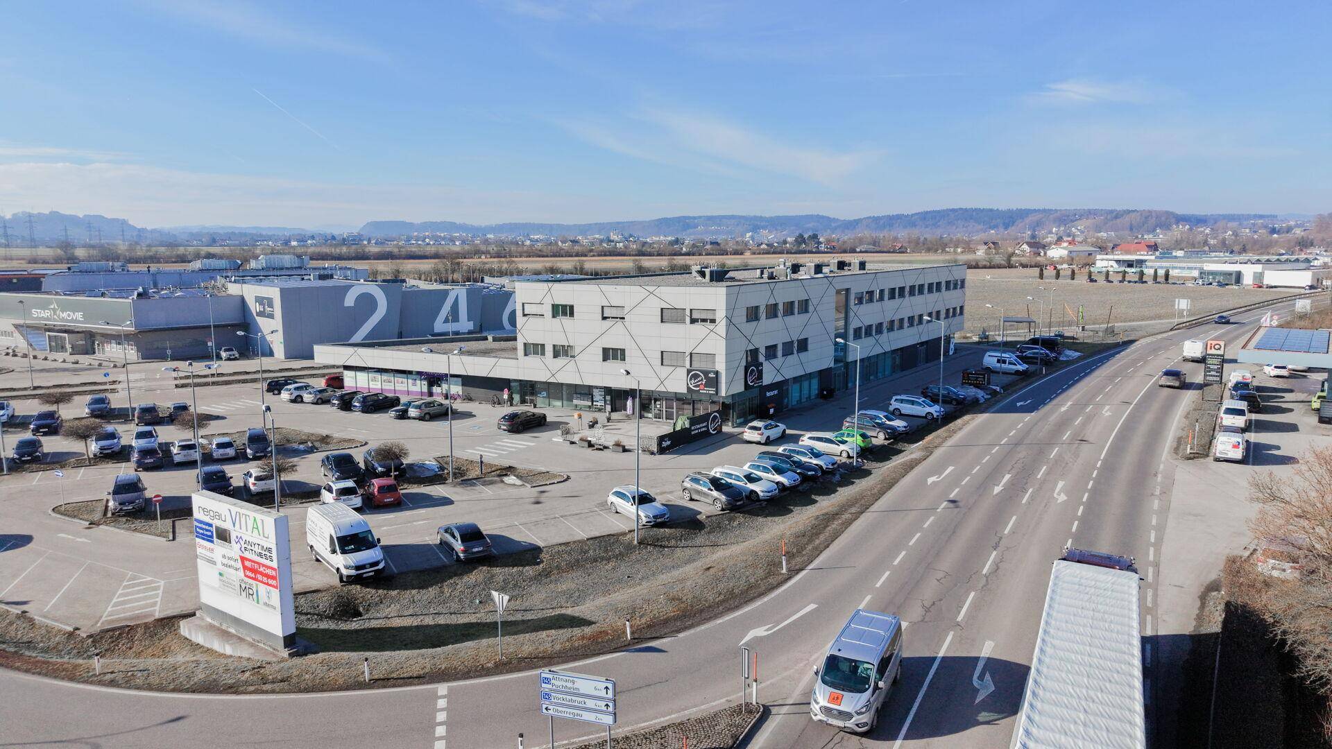 Luftaufnahme eines Gewerbegebiets mit mehreren Gebäuden, Parkplätzen und einer belebten Straße unter blauem Himmel.