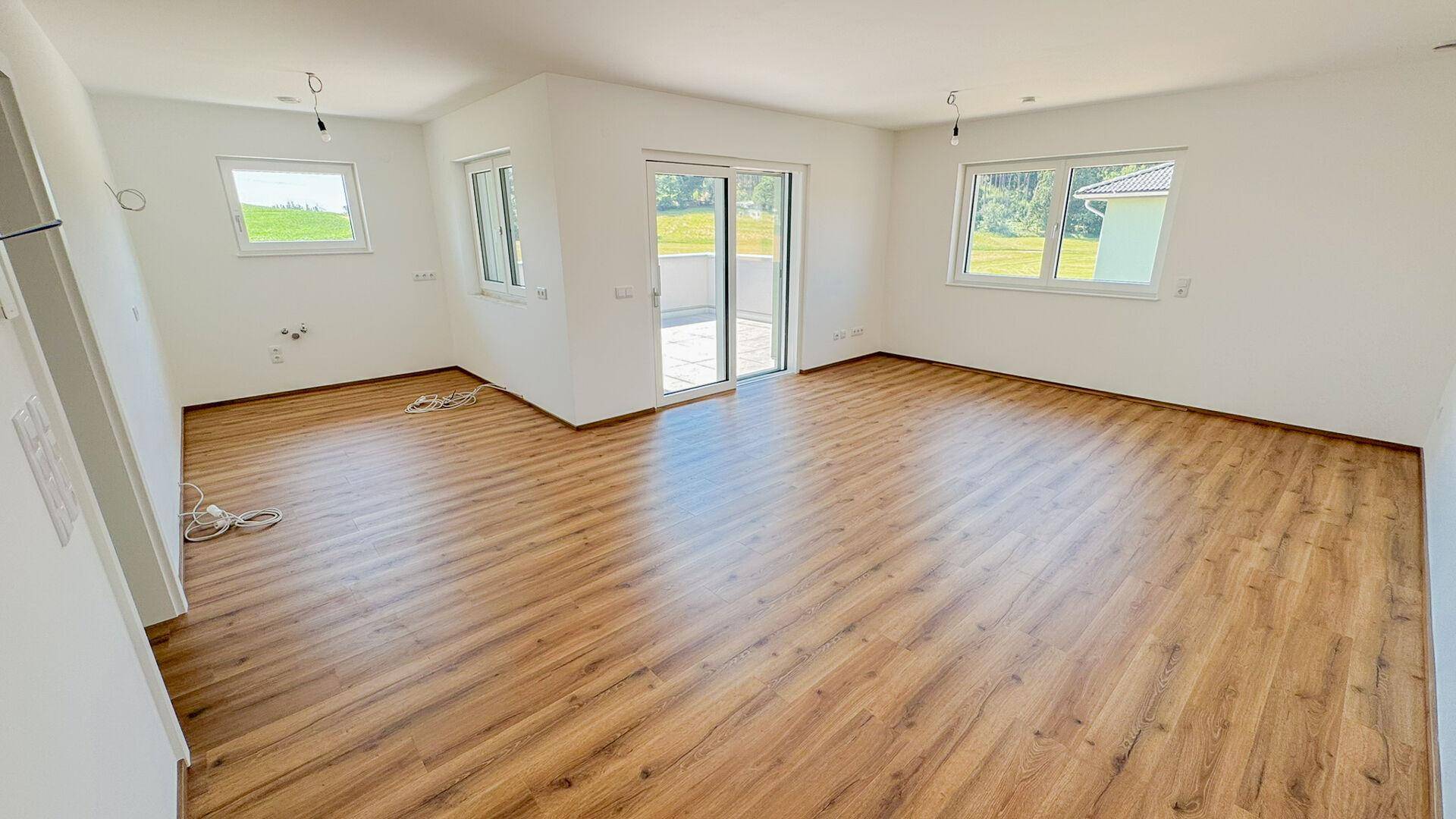 Geräumiges, leeres Wohnzimmer mit Laminatboden, großen Fenstern und Zugang zur Terrasse.