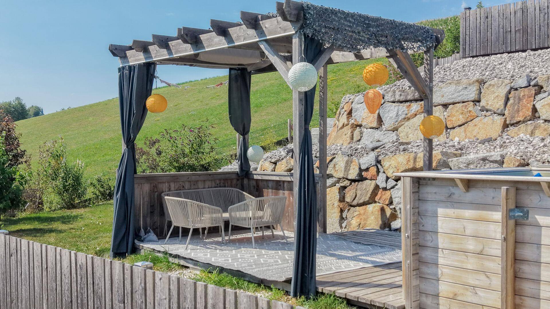 Gemütlicher Außenbereich mit Pergola, Sitzgelegenheiten und Whirlpool auf einer Holzterrasse mit Blick ins Grüne.