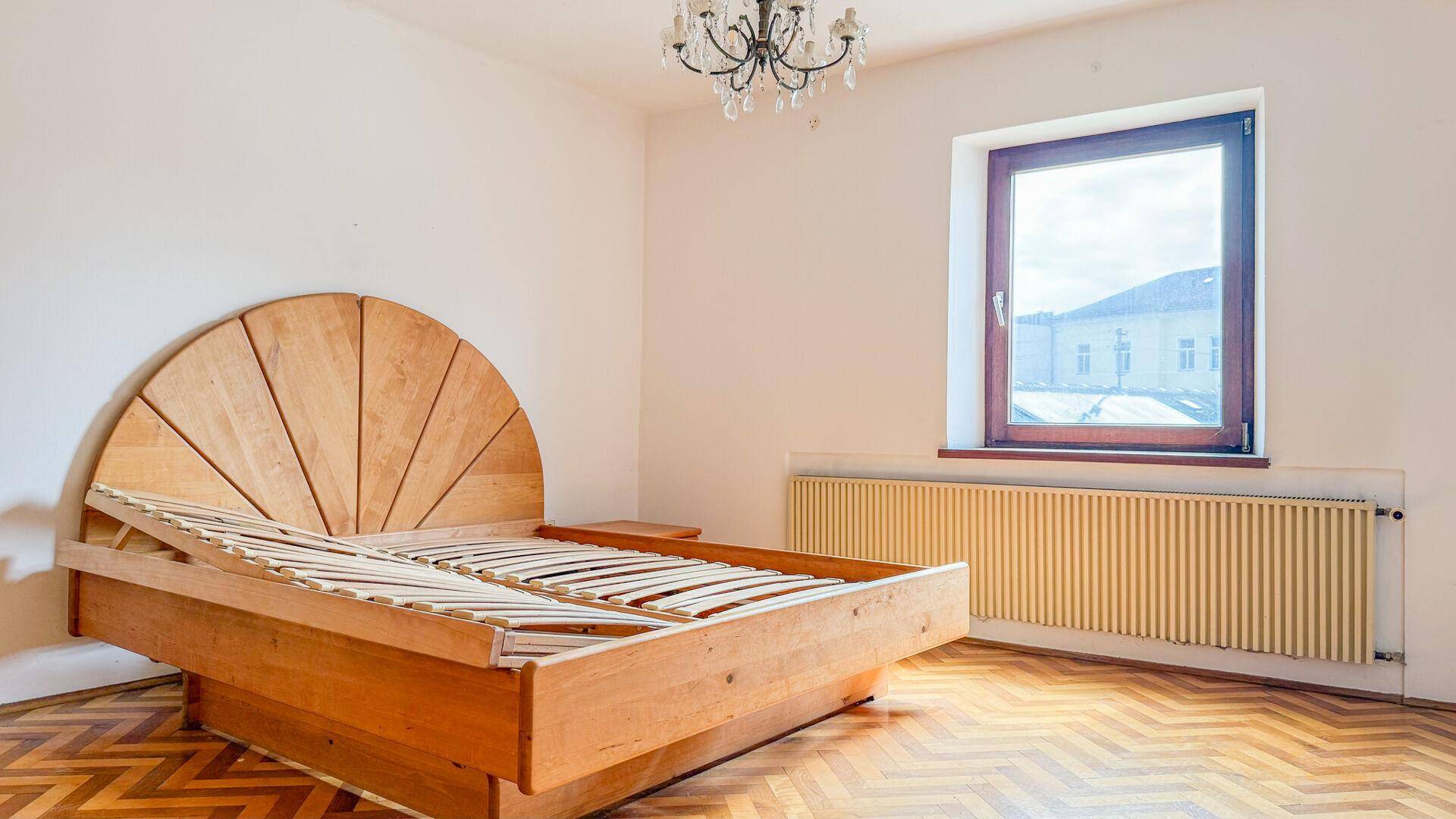Schlafzimmer mit Fischgrätparkett, Holz-Bettrahmen, Fenster und einem Kronleuchter.