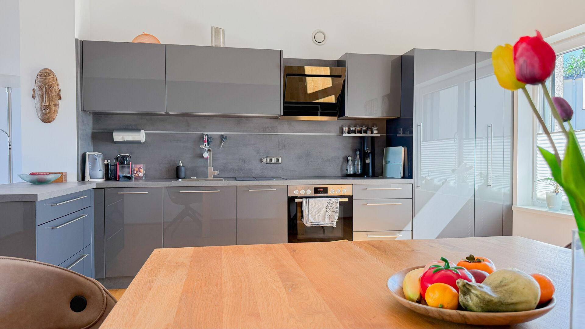 Moderne Küche mit grauen Hochglanzfronten, integriertem Backofen und Esstisch für gemeinsame Mahlzeiten.