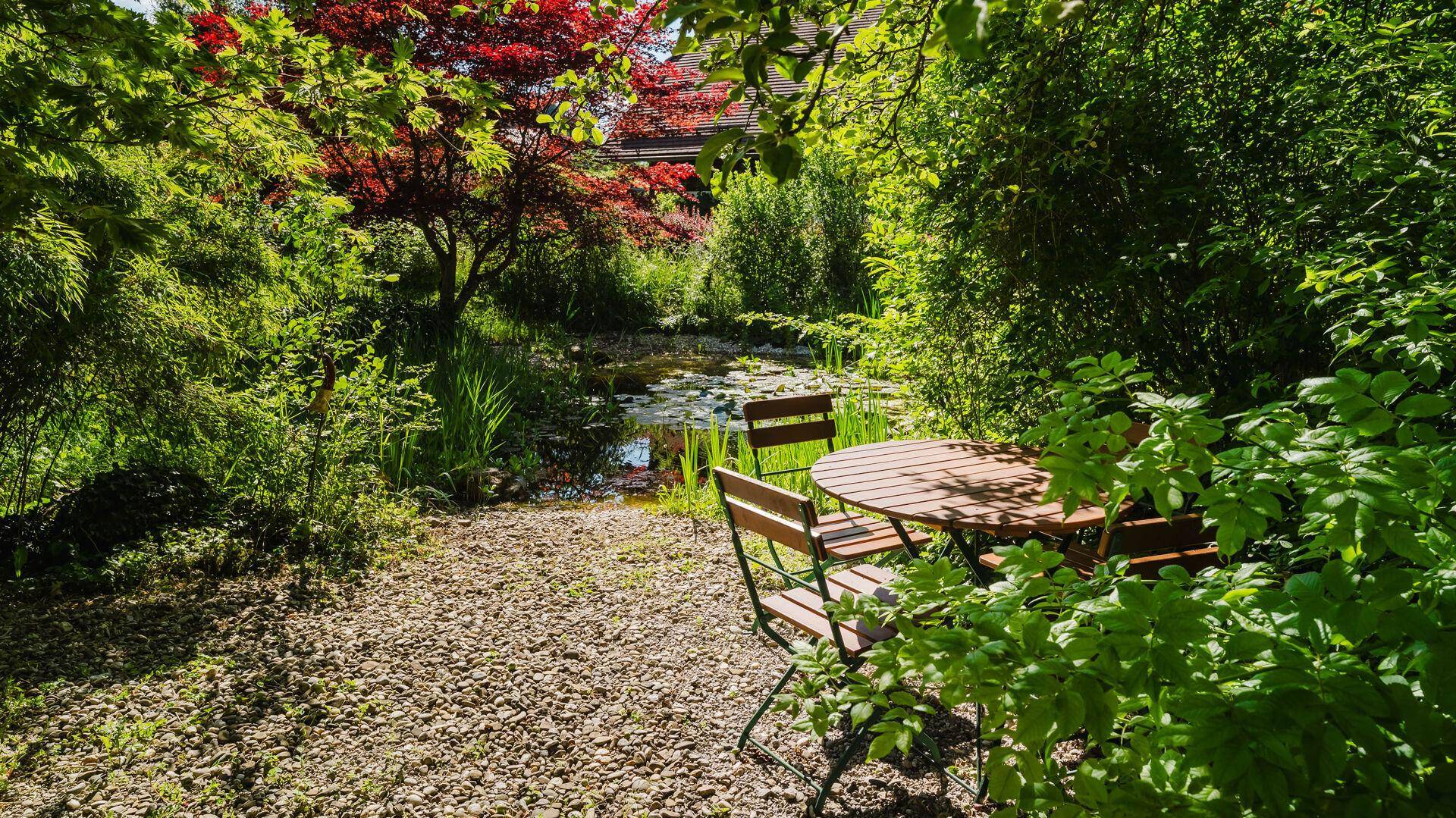 Idyllischer Gartenbereich mit Teich, Sitzgelegenheiten und üppiger Vegetation.