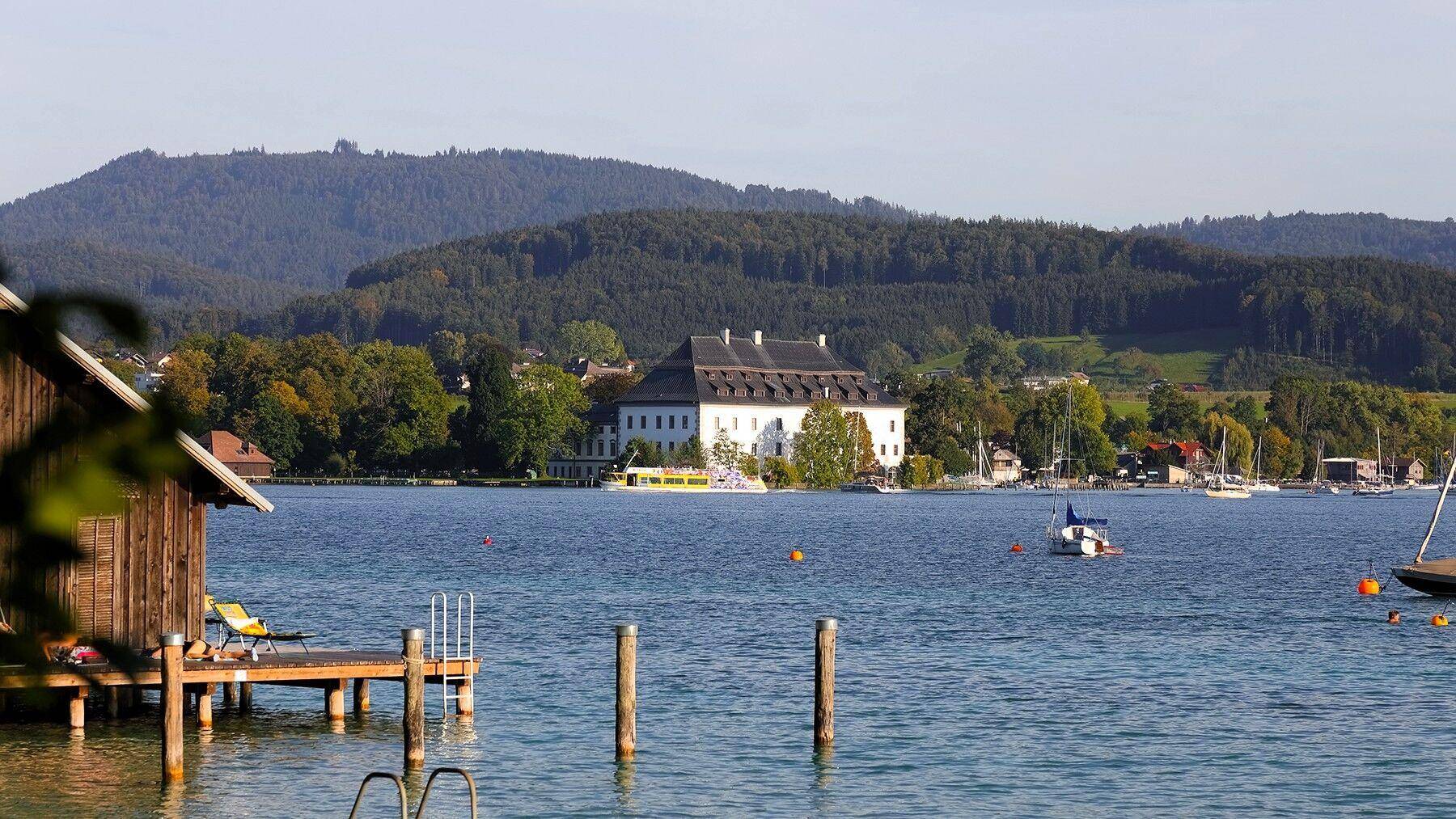 Malerischer Seeblick mit einem historischen Gebäude am Ufer, umgeben von Bäumen und Bergen.