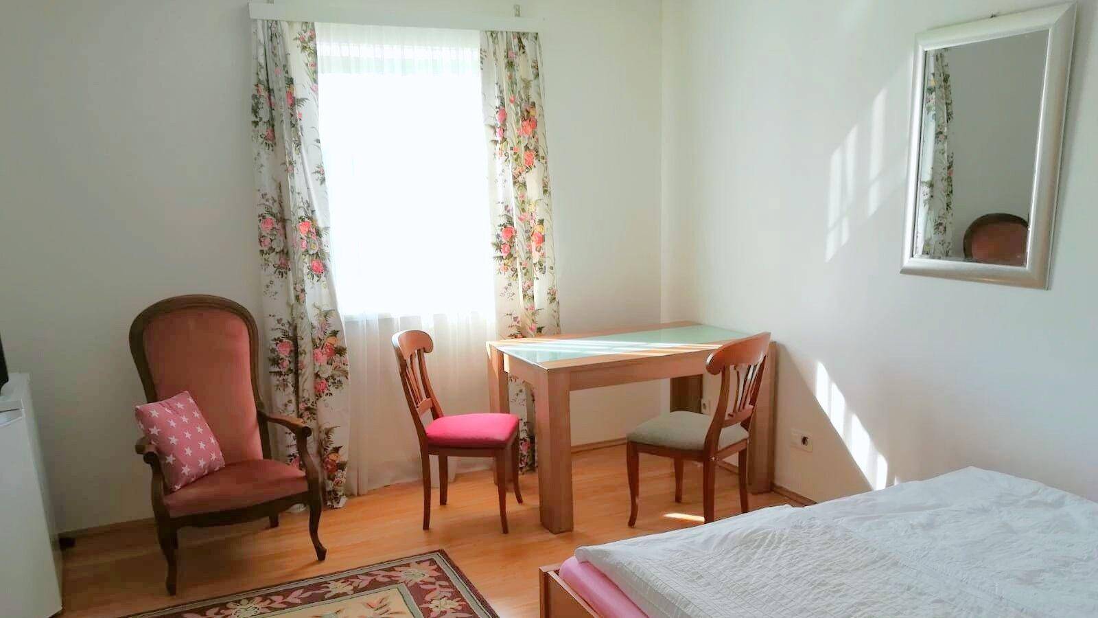 Helles Schlafzimmer mit Doppelbett, Esstisch und floralen Vorhängen.