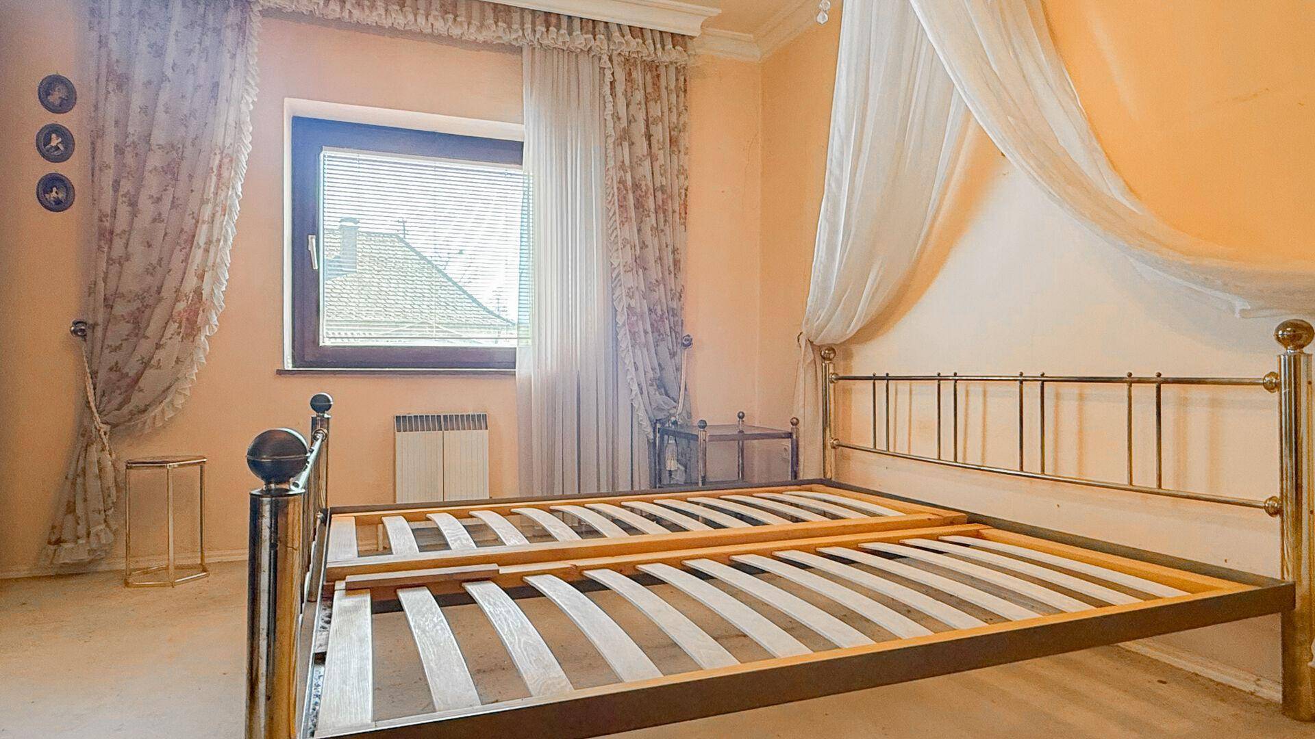 Schlafzimmer mit Metallbettgestell und Vorhängen, gut belichtet durch ein Fenster.