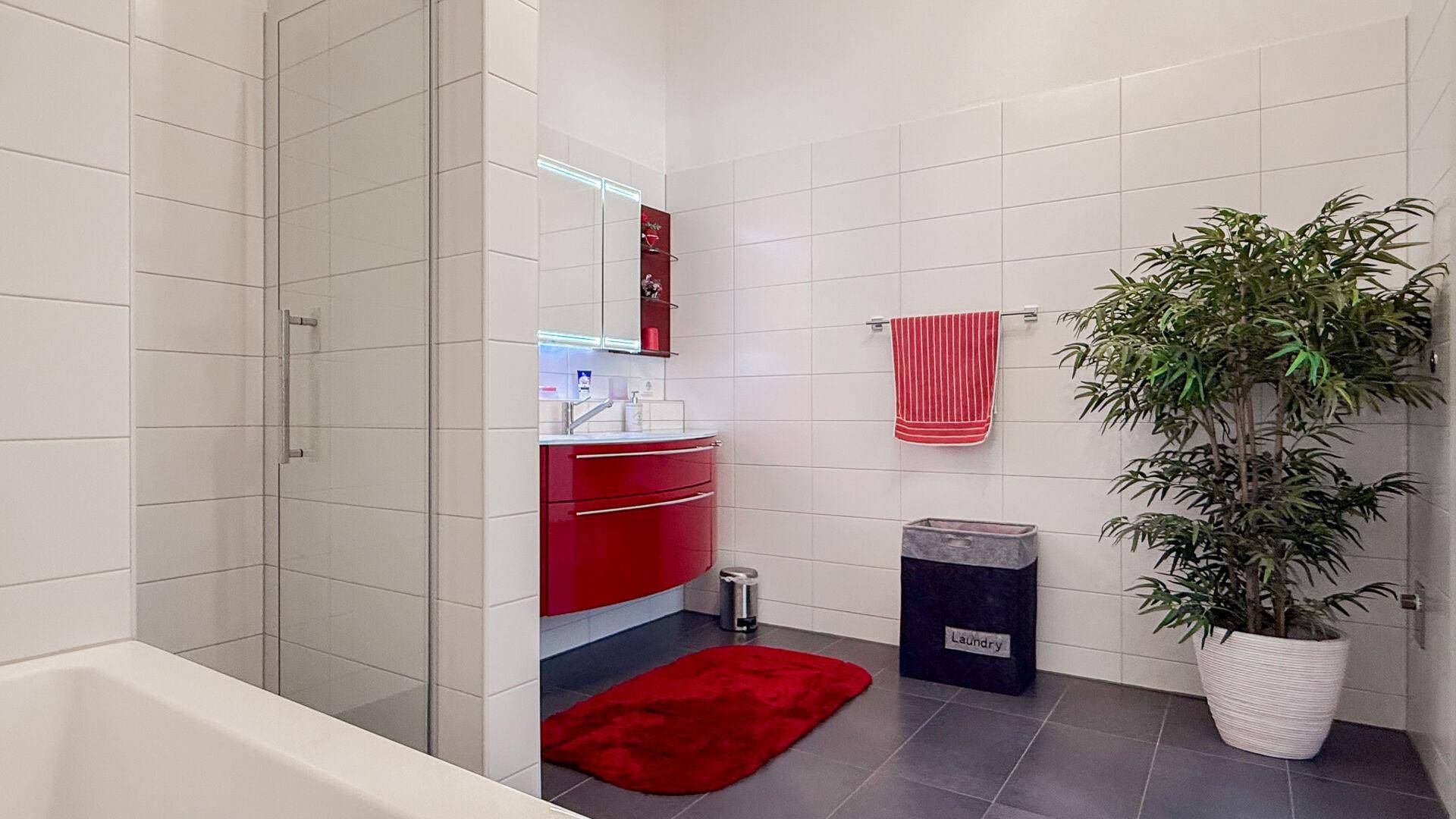 Modernes Badezimmer mit weißen Fliesen, rotem Waschtisch, Badewanne und Duschbereich.