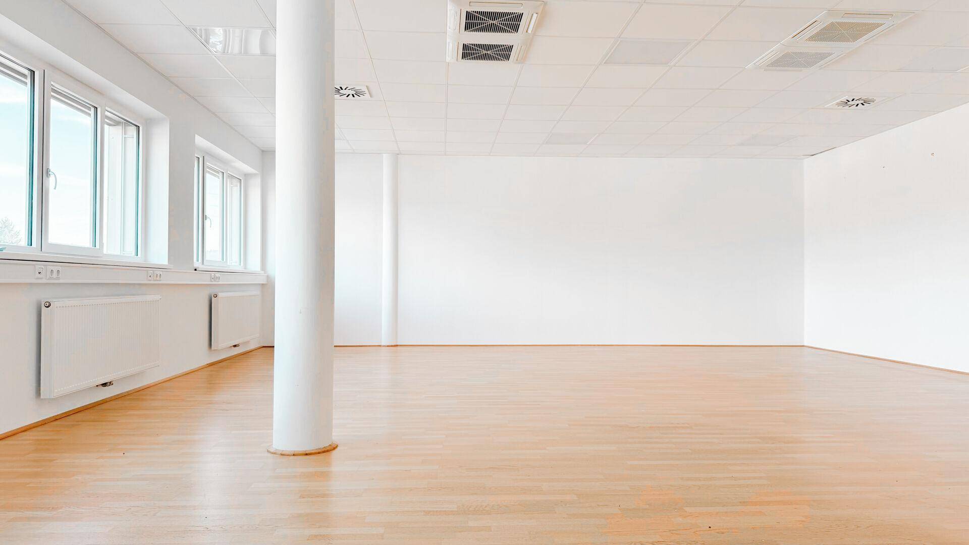 Geräumiger, heller Büroraum mit großen Fenstern, einer Säule und glänzendem Holzfußboden.