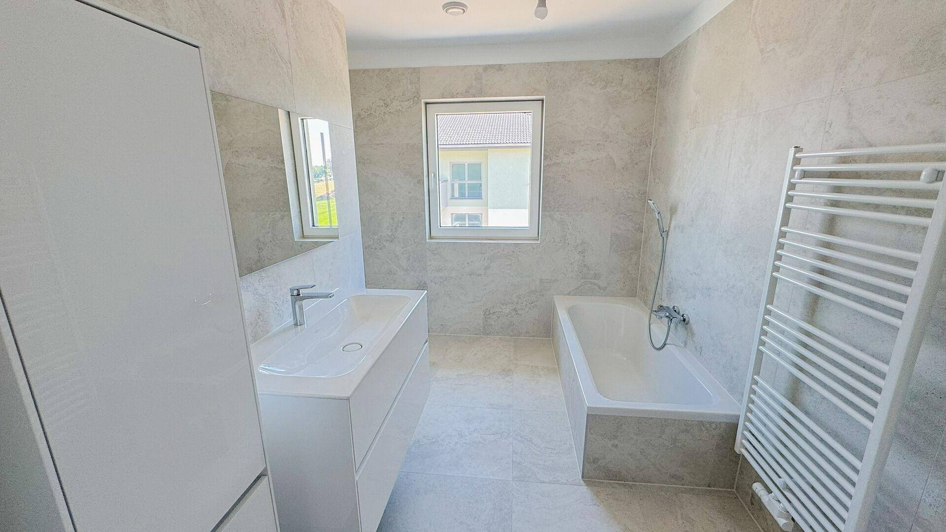 Modernes Badezimmer mit hellgrauen Fliesen, Badewanne, Einzelwaschbecken und beheiztem Handtuchhalter.