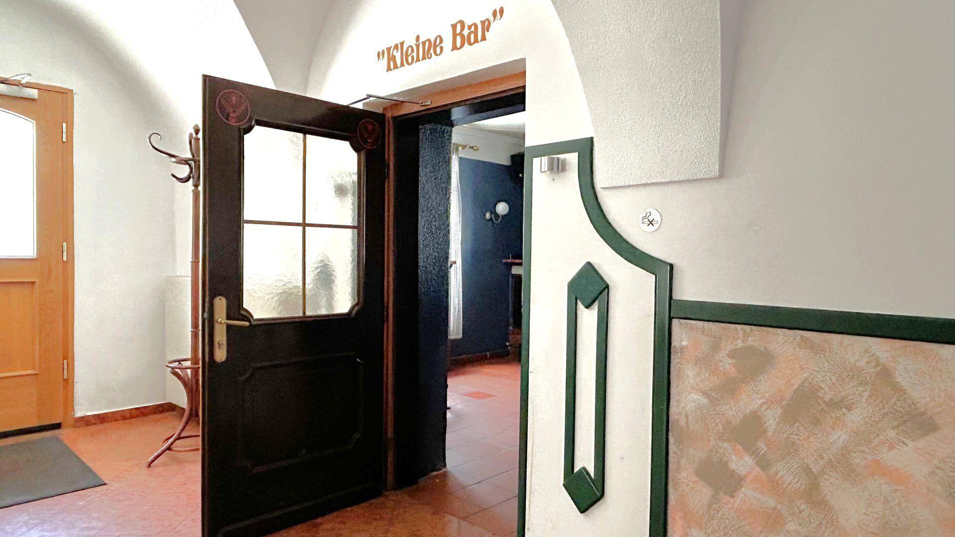 Blick in einen Flur mit gewölbter Decke und einer Tür, die zu einer "Kleinen Bar" führt.