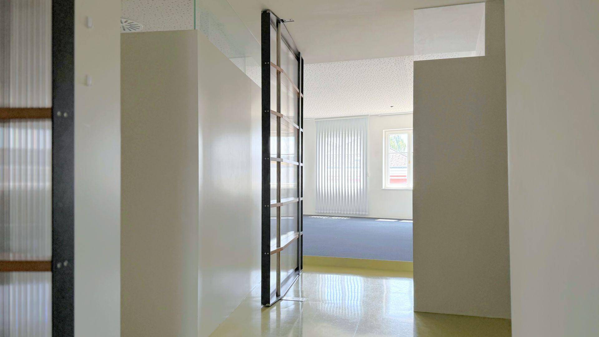 Heller Übergangsbereich mit glänzendem Boden und Blick in einen weiteren Raum mit vertikalen Jalousien an den Fenstern.