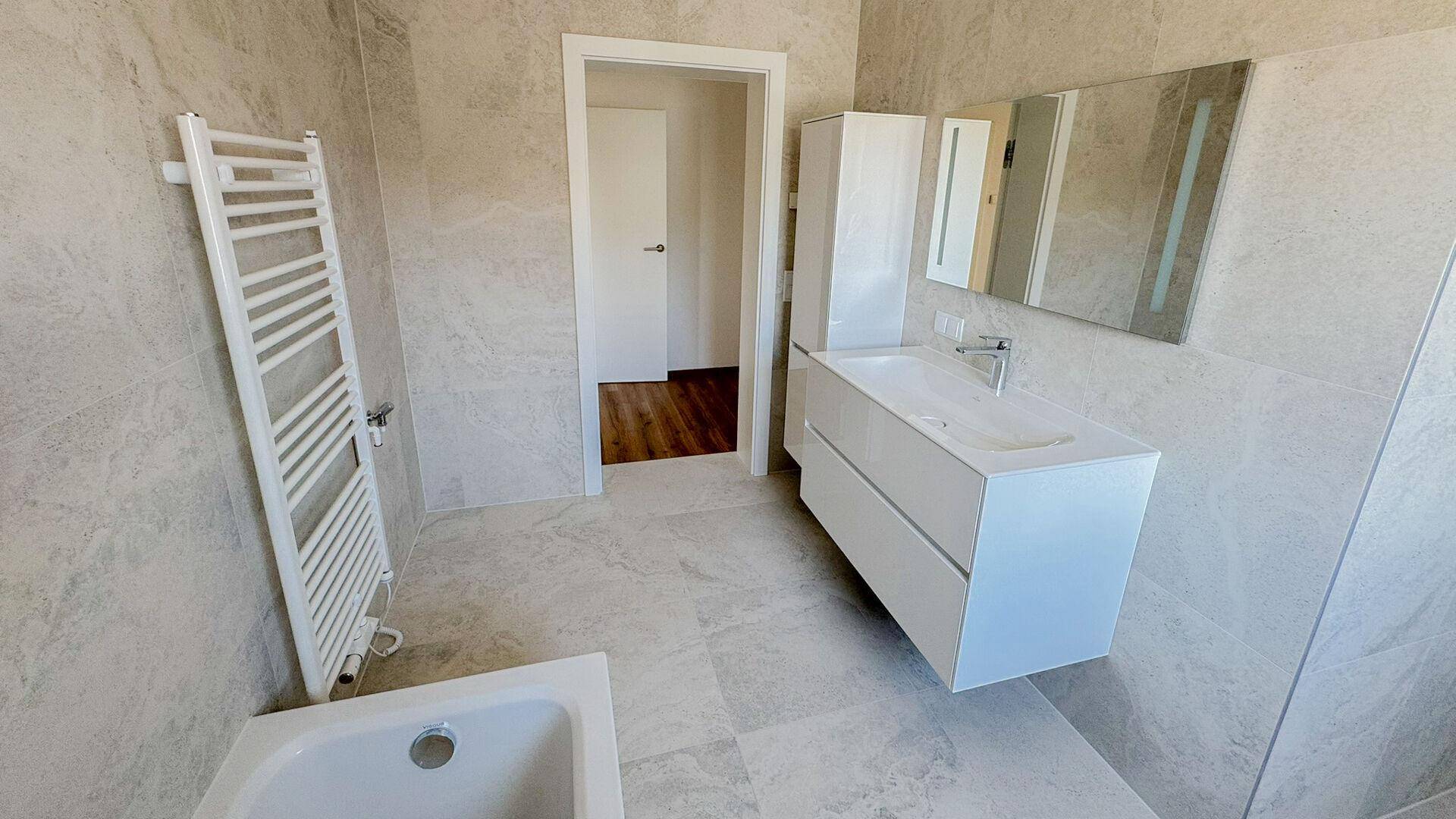 Modernes Badezimmer mit hellgrauen Fliesen, Waschtisch, Spiegel und beheiztem Handtuchhalter.