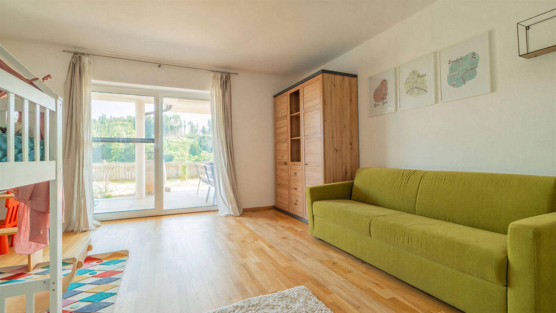 Geräumiges Zimmer mit Holzboden, grünem Sofa und großem Fenster mit Blick ins Grüne.