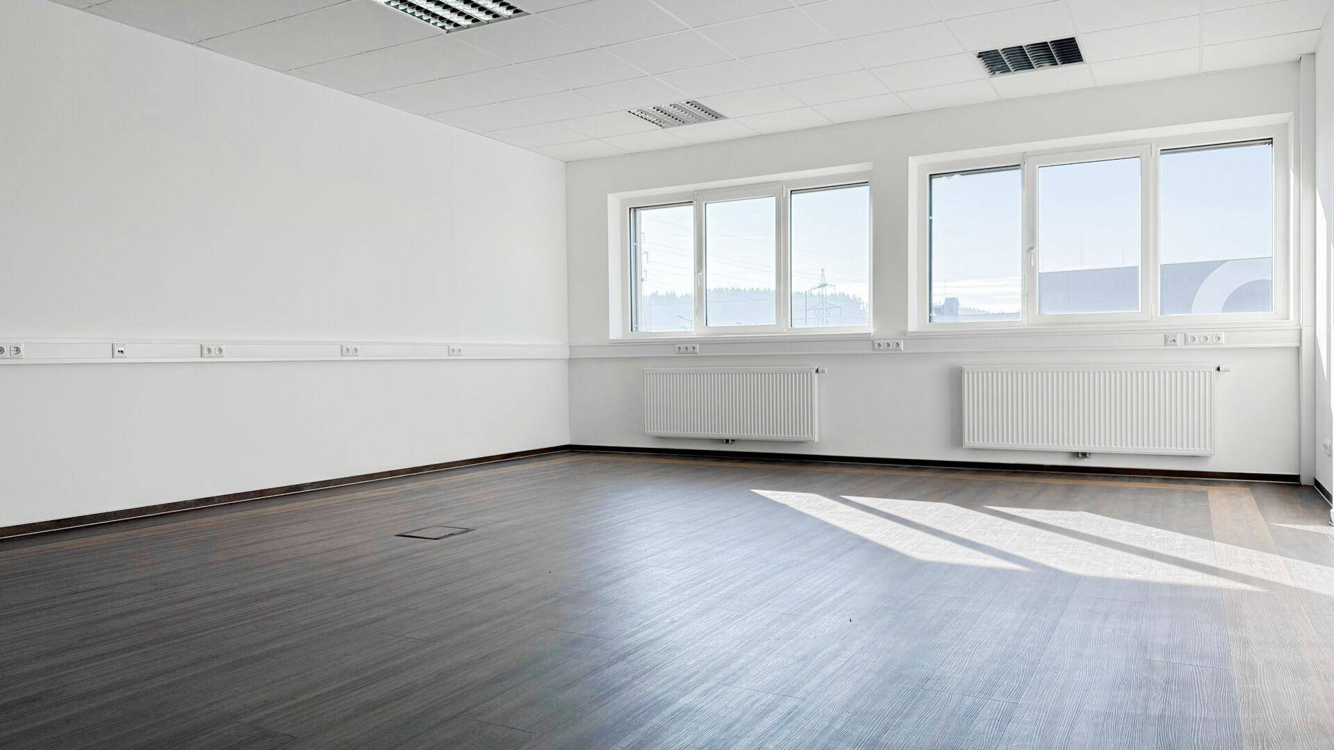 Großer, lichtdurchfluteter Büroraum mit mehreren Fenstern und dunklem Boden, bietet viel Platz.