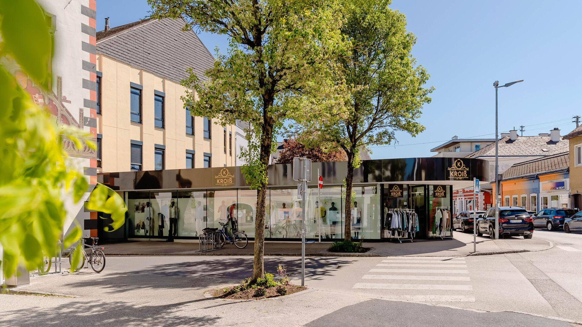 Moderne Geschäftsfassade mit großen Schaufenstern und Bäumen an einer städtischen Kreuzung.