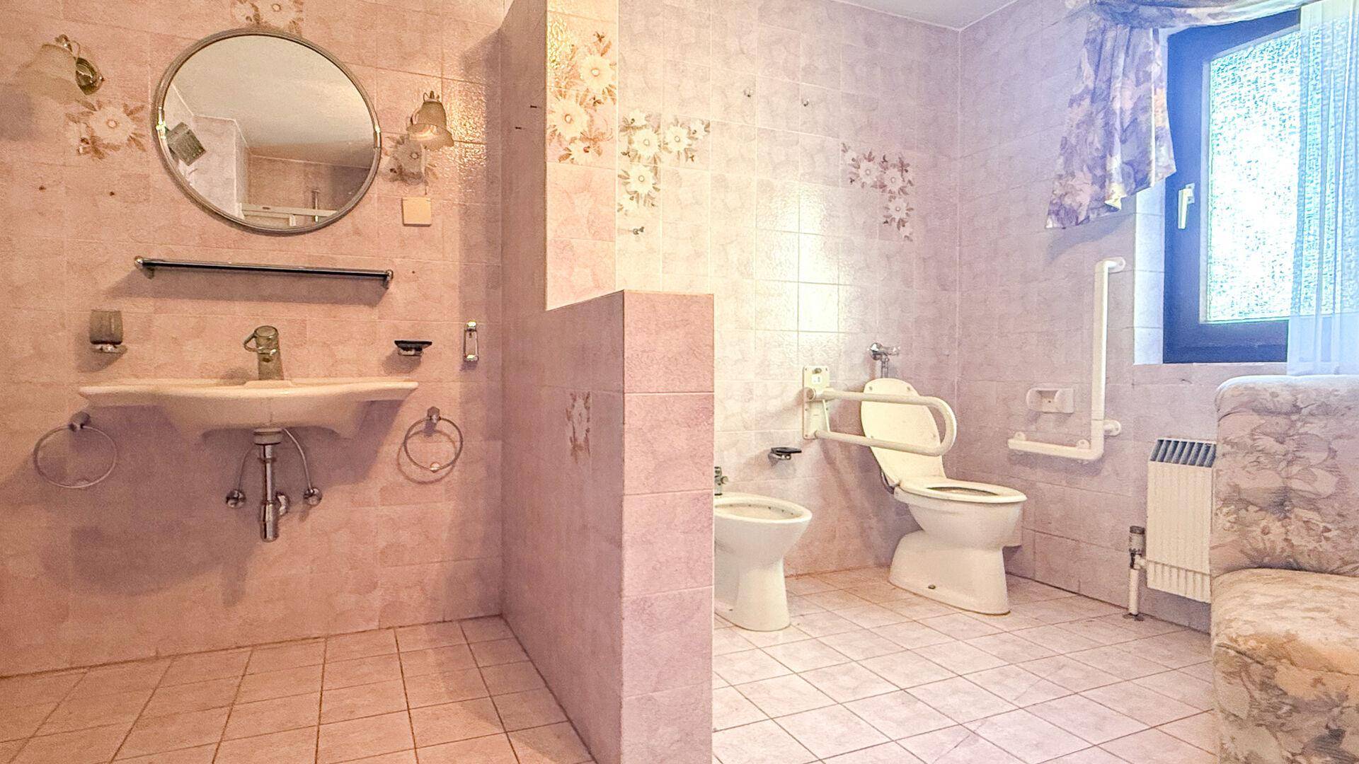 Geräumiges Badezimmer mit rosa Fliesen, Waschbecken und WC mit Haltegriffen am Fenster.