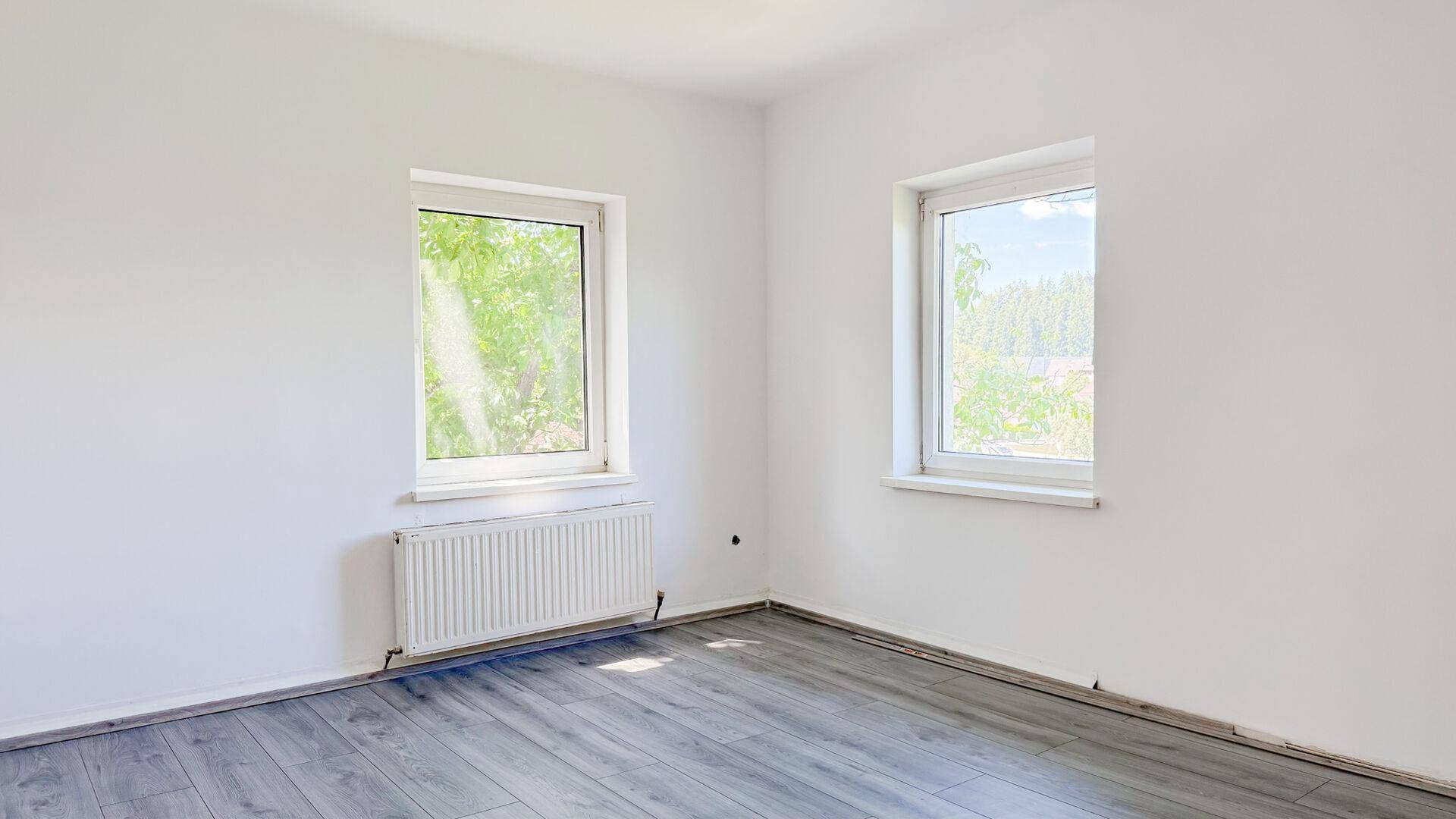 Freundlicher Raum mit weiß gestrichenen Wänden, grauem Laminatboden und zwei Fenstern mit grüner Aussicht.