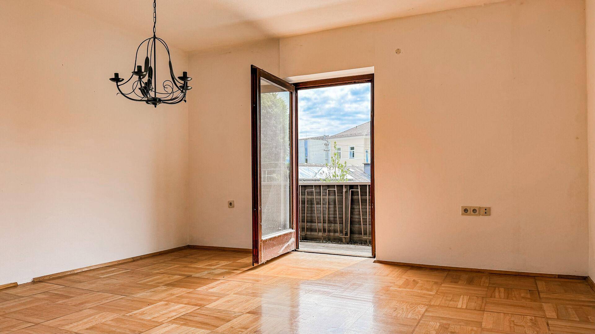 Zimmer mit Parkettboden, Kronleuchter und Zugang zu einem kleinen Balkon mit Blick ins Freie.