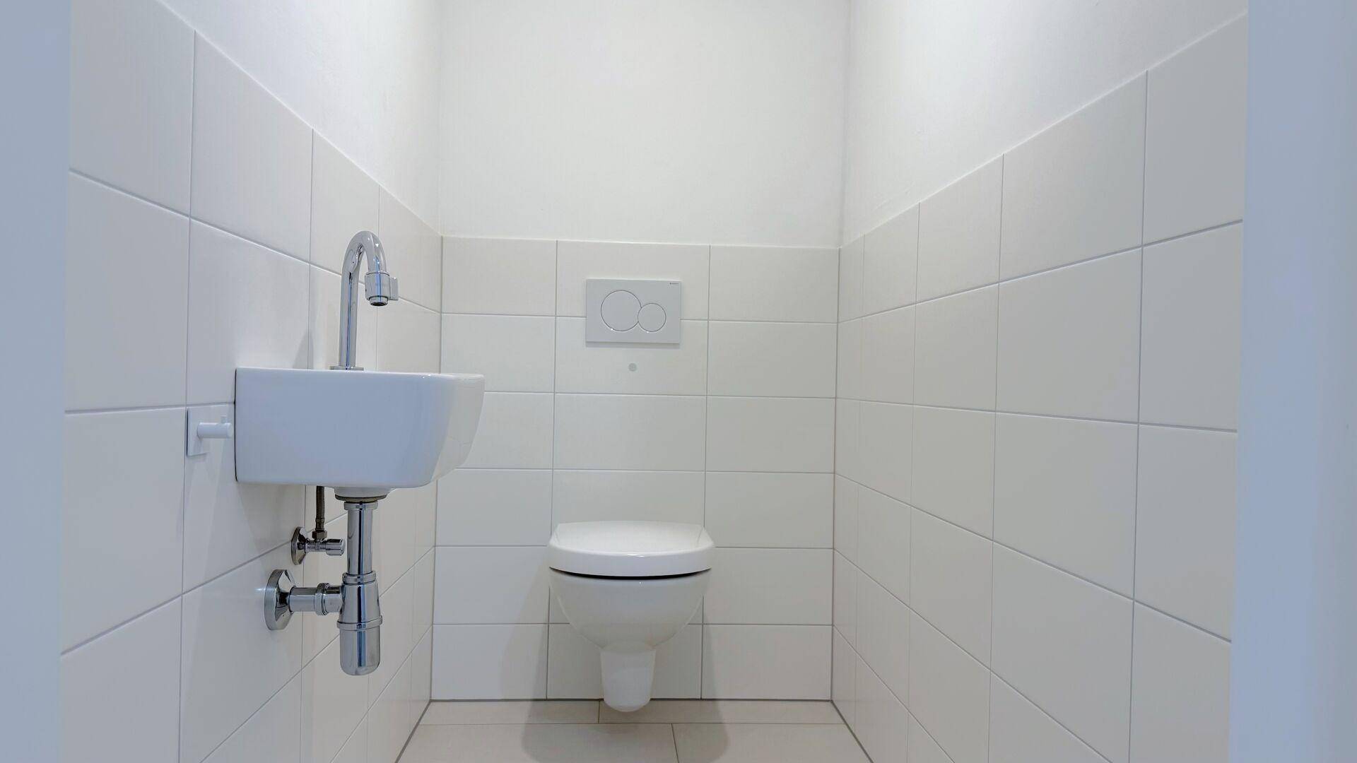 Separates WC mit weißen Fliesen, wandhängender Toilette und einem kleinen Handwaschbecken.